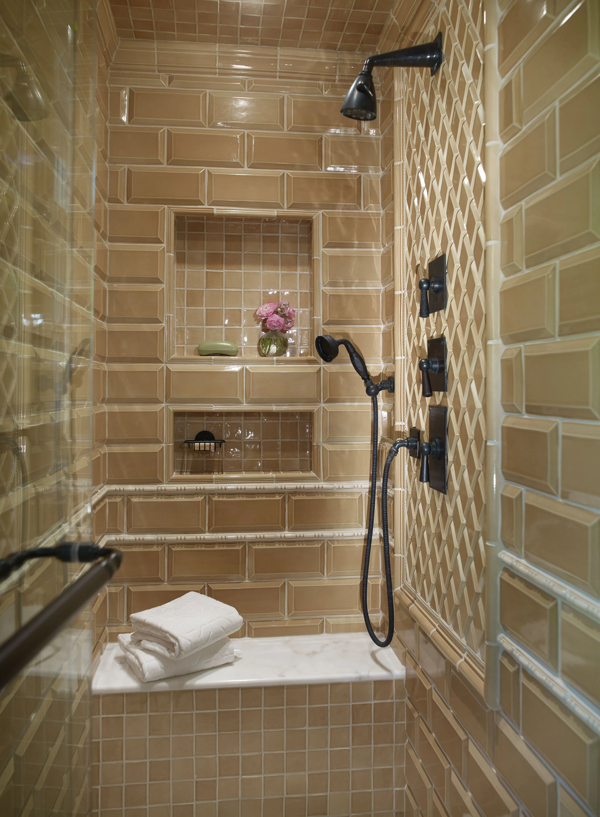 Inside a shower with tan tile walls, a built-in niche with pink flowers and a soap, a marble seat with folded towels, and multiple black shower fixtures, including a showerhead and handheld shower. Interior design by Sarah Barnard. 