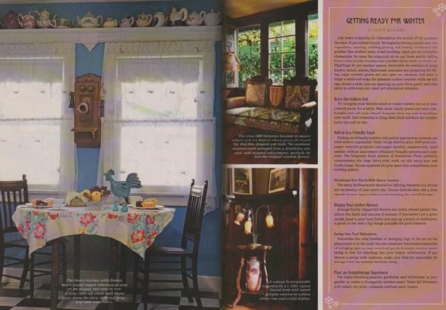 Kitchen with a blue wall, window, and a dining table covered with a floral tablecloth, set with dishes and a tea set. Chairs are around the table, and decorative china are displayed on a shelf above.