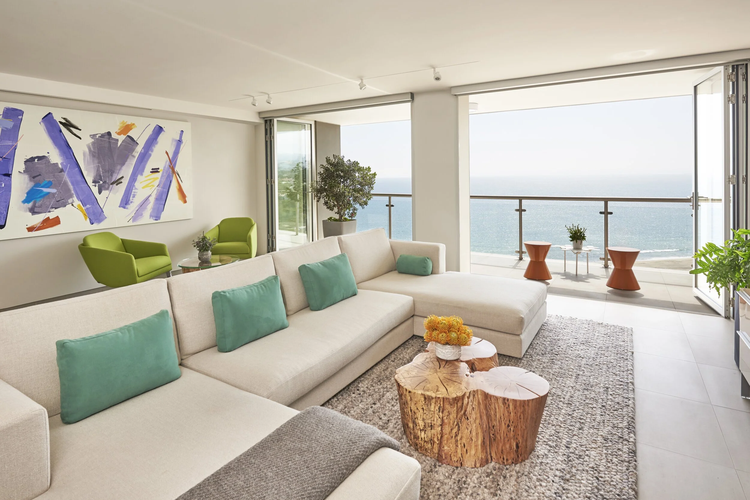 Living room with large beige sectional sofa, blue throw pillows, wooden coffee table with flowers, green armchairs, large art piece on white wall, potted plants, and a balcony with ocean view. Interior design by Sarah Barnard. 