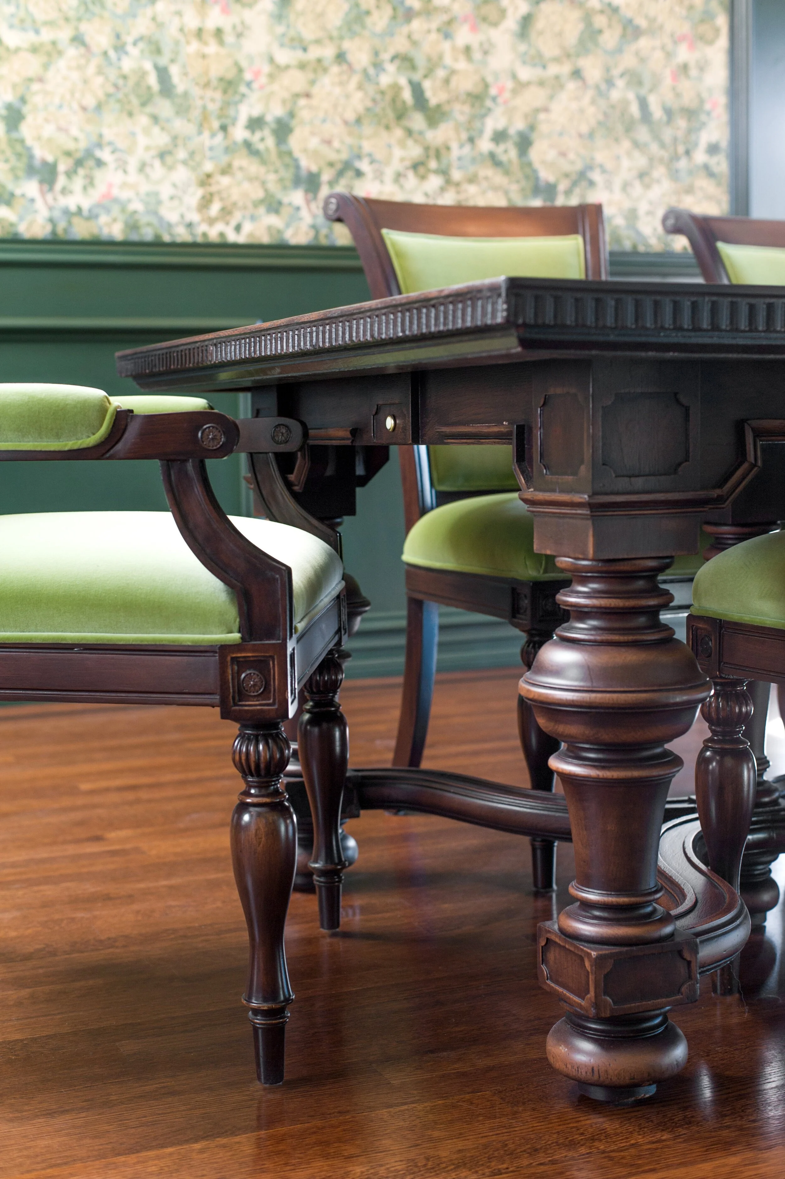 A close-up view of a wooden dining table with carved legs, surrounded by chairs with green upholstered seats, set against a wall with a patterned wallpaper. Interior design by Sarah Barnard. 
