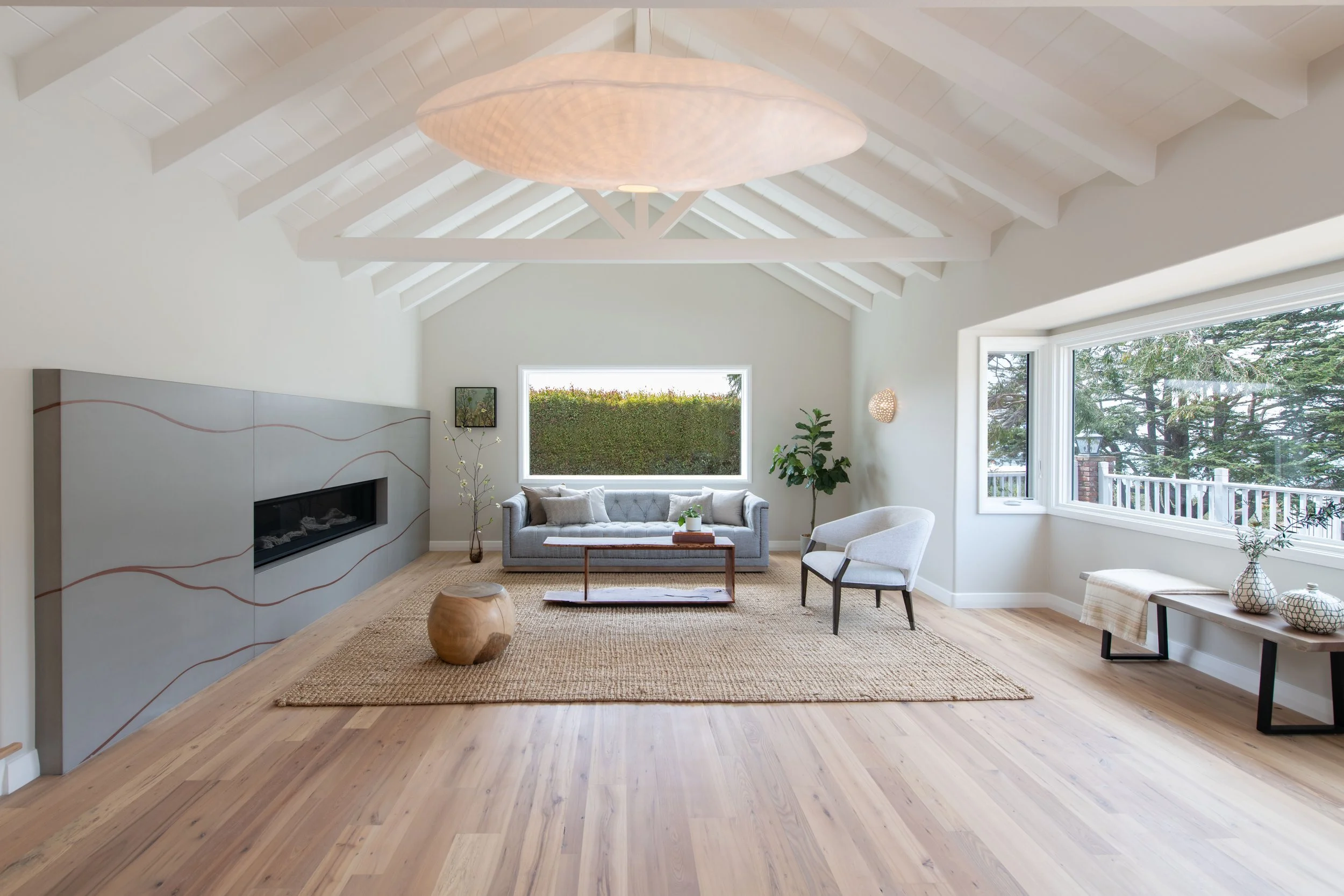 Bright living room with a high, white vaulted ceiling, large window view of trees, light gray sofa, white armchair, small wooden coffee table, and decorative items, with a woven rug on light hardwood floor. Interior design by Sarah Barnard. 