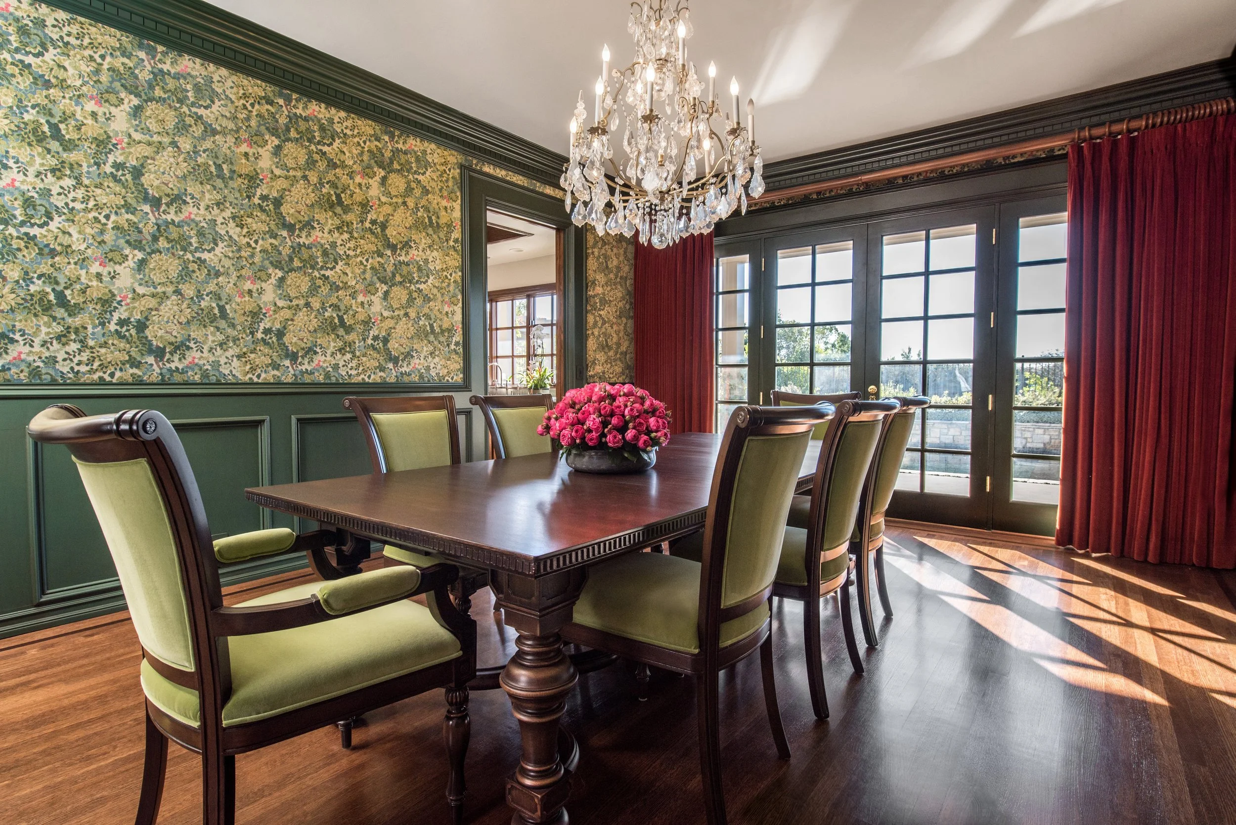 Elegant dining room with a large wooden table, six green upholstered chairs, a chandelier, floral wallpaper, red curtains, and large glass doors leading outside. Interior design by Sarah Barnard.
