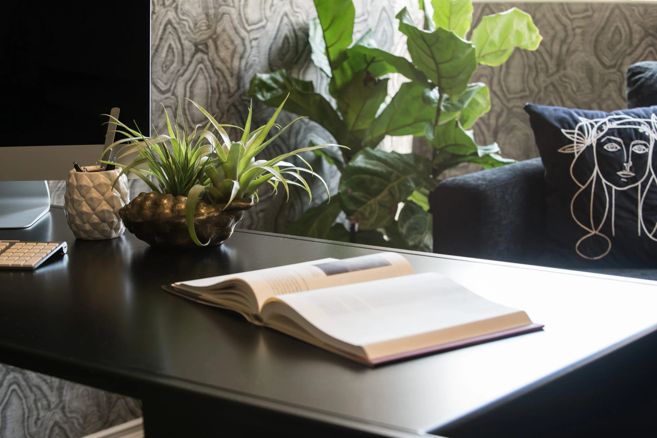 Black desk with an open book, small plants, and a computer against patterned wallpaper. Interior design by Sarah Barnard Design Manhattan Beach.