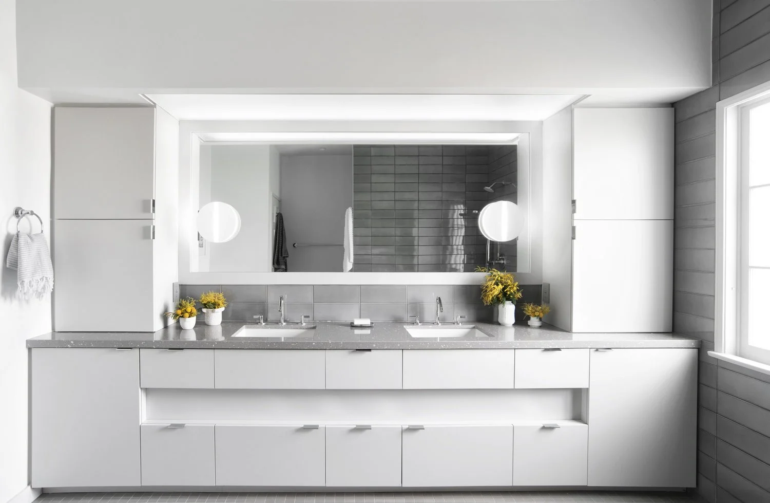 Minimal white bathroom with double sinks, large mirror, and small yellow flowers.