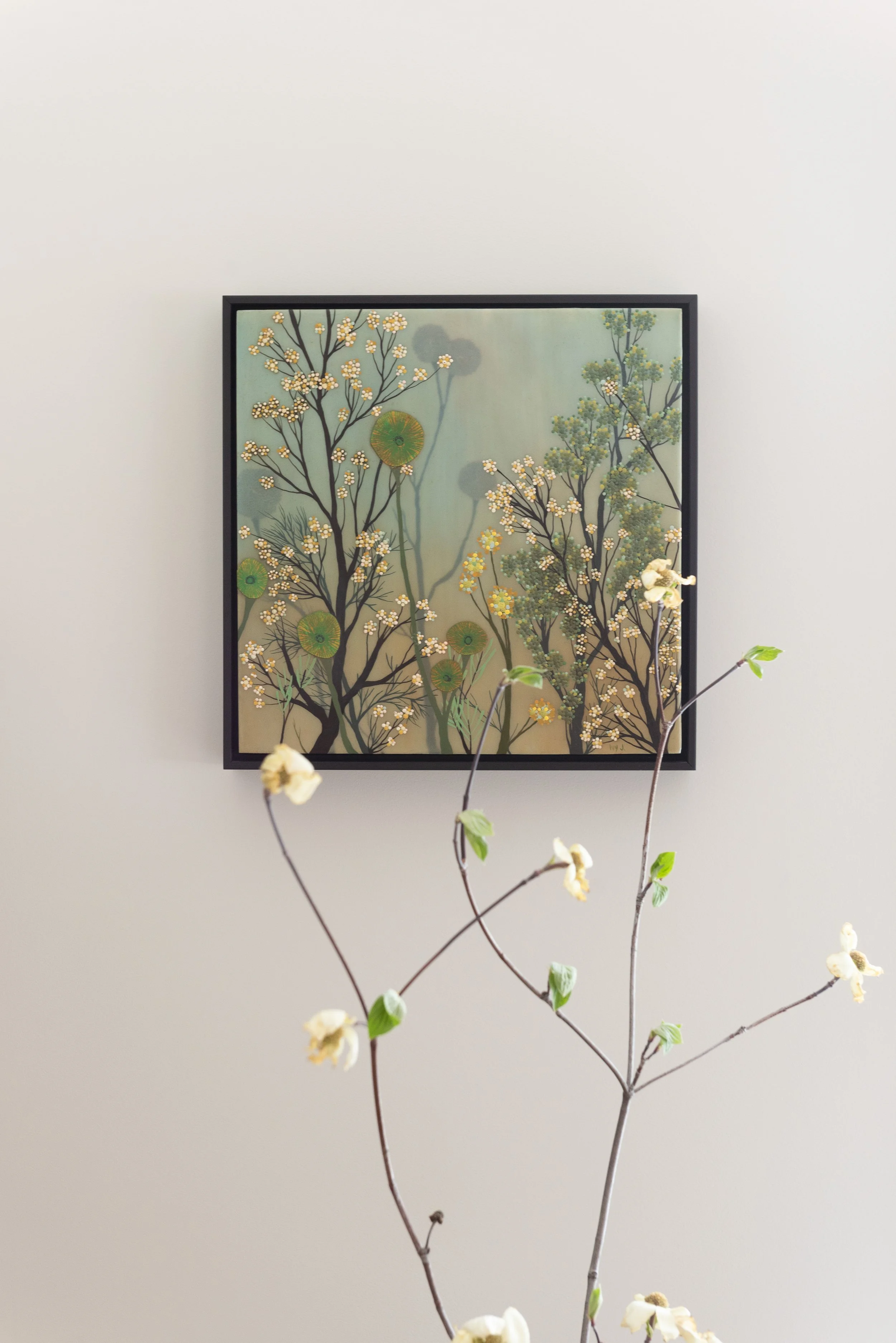 Frame painting of trees with white flowers and green round seed pods, hanging on a white wall, with a branch of cream-colored flowers in front. Interior design by Sarah Barnard. 