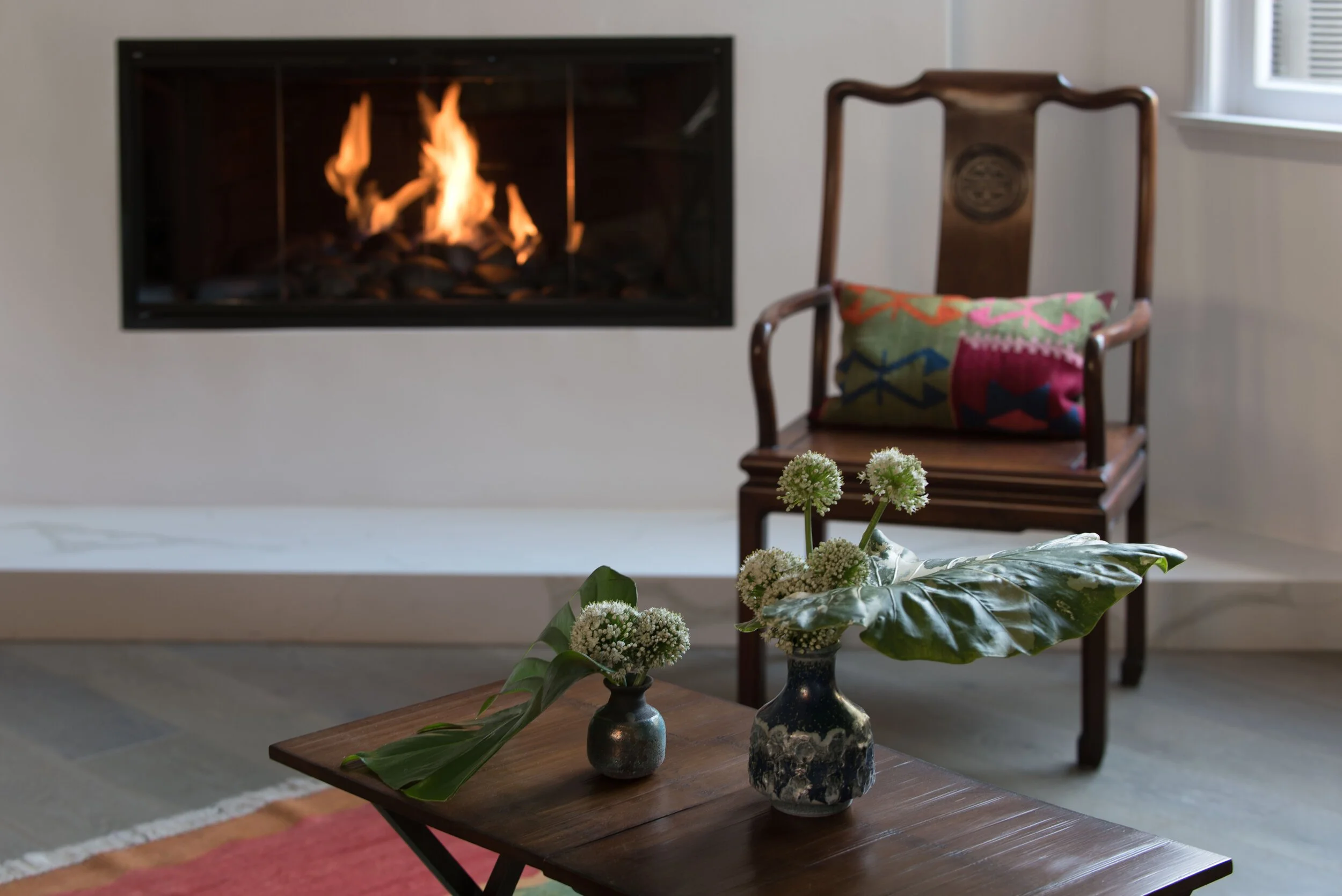 Living room with a lit fireplace, a wooden chair with a colorful pillow, and a wooden coffee table with two vases of flowers. Interior design by Sarah Barnard. 