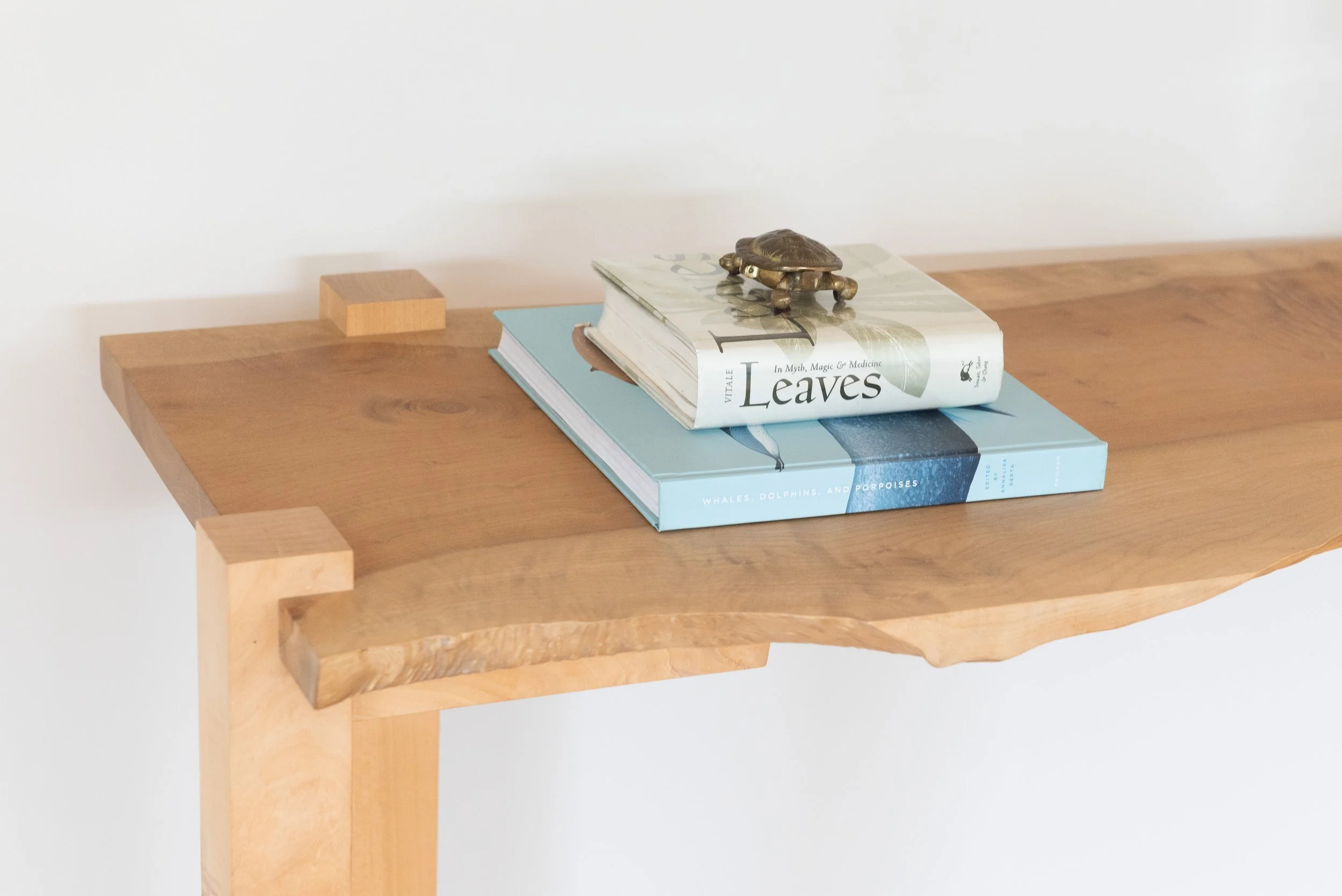 Wooden table with a stack of two books, the top book titled 'Leaves' and 'In Myth, Magic & Medicine,' with a small decorative turtle figurine on top. Interior design by Sarah Barnard. 