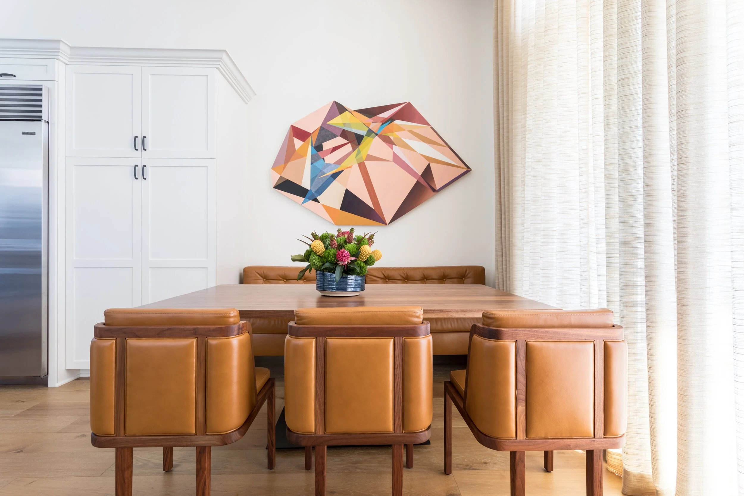 A dining room with a wooden table surrounded by six tan upholstered chairs. A blue vase with a colorful flower arrangement sits on the table. A geometric colorful wall art hangs above the table. Interior design by Sarah Barnard. 