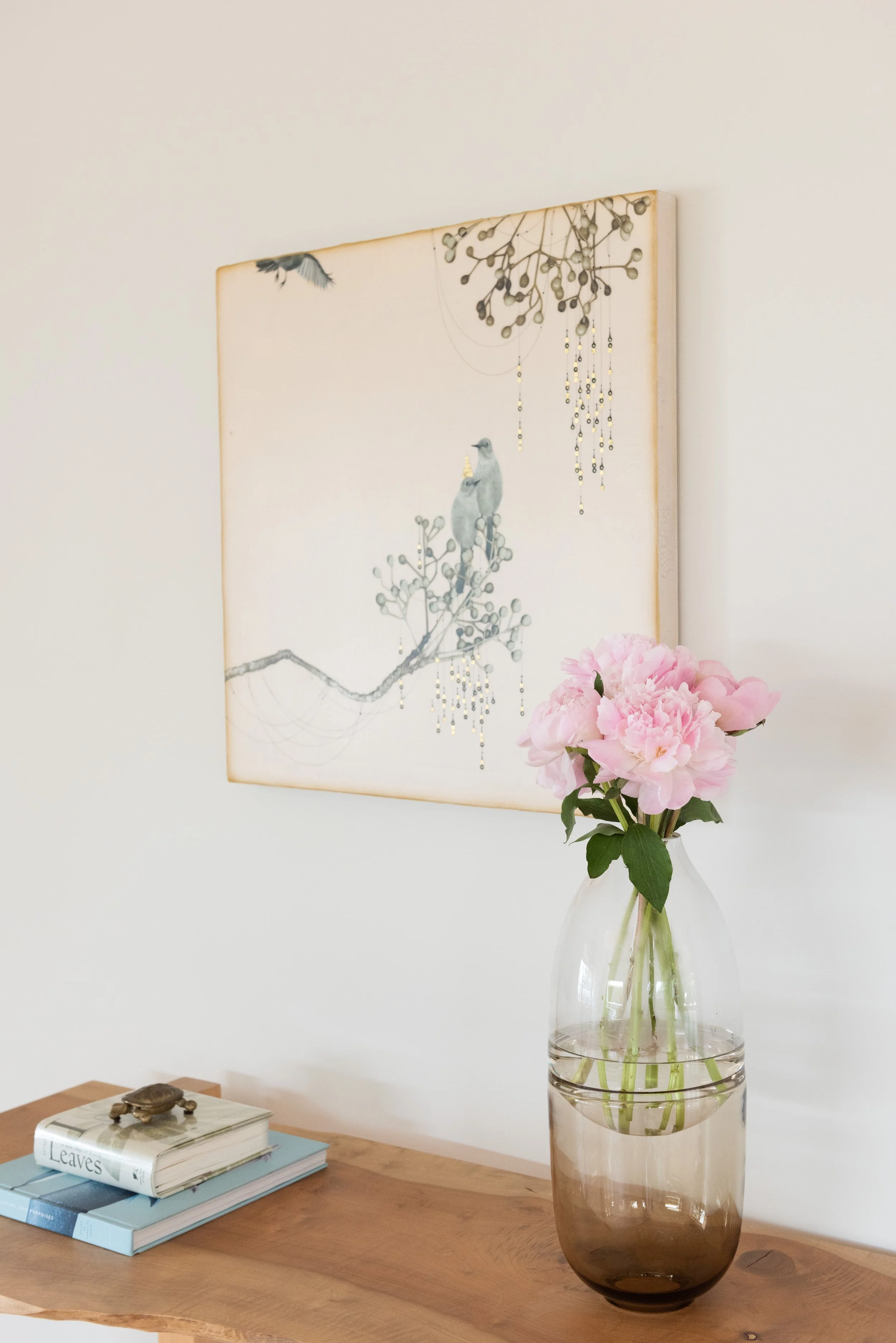 A clear glass vase with pink peonies on a wooden table, with books and a small decorative turtle on top. A framed artwork featuring birds and branches hangs on a cream wall in the background. Interior design by Sarah Barnard. 
