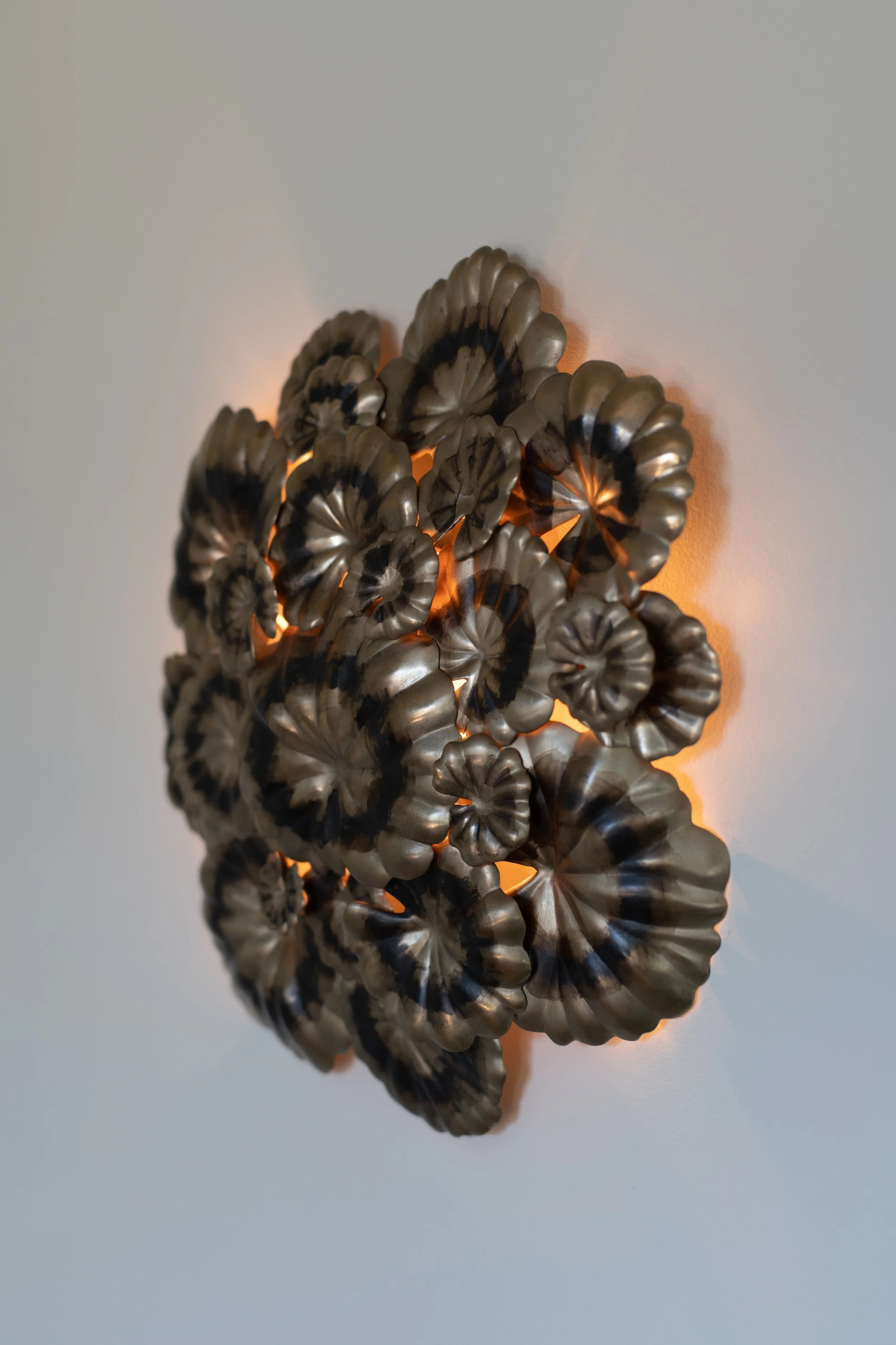 Wall-mounted Geranium decorative sculpture composed of metallic, spiral-shaped elements with a black and brass finish, illuminated from behind to create a glowing effect. Interior design by Sarah Barnard. 