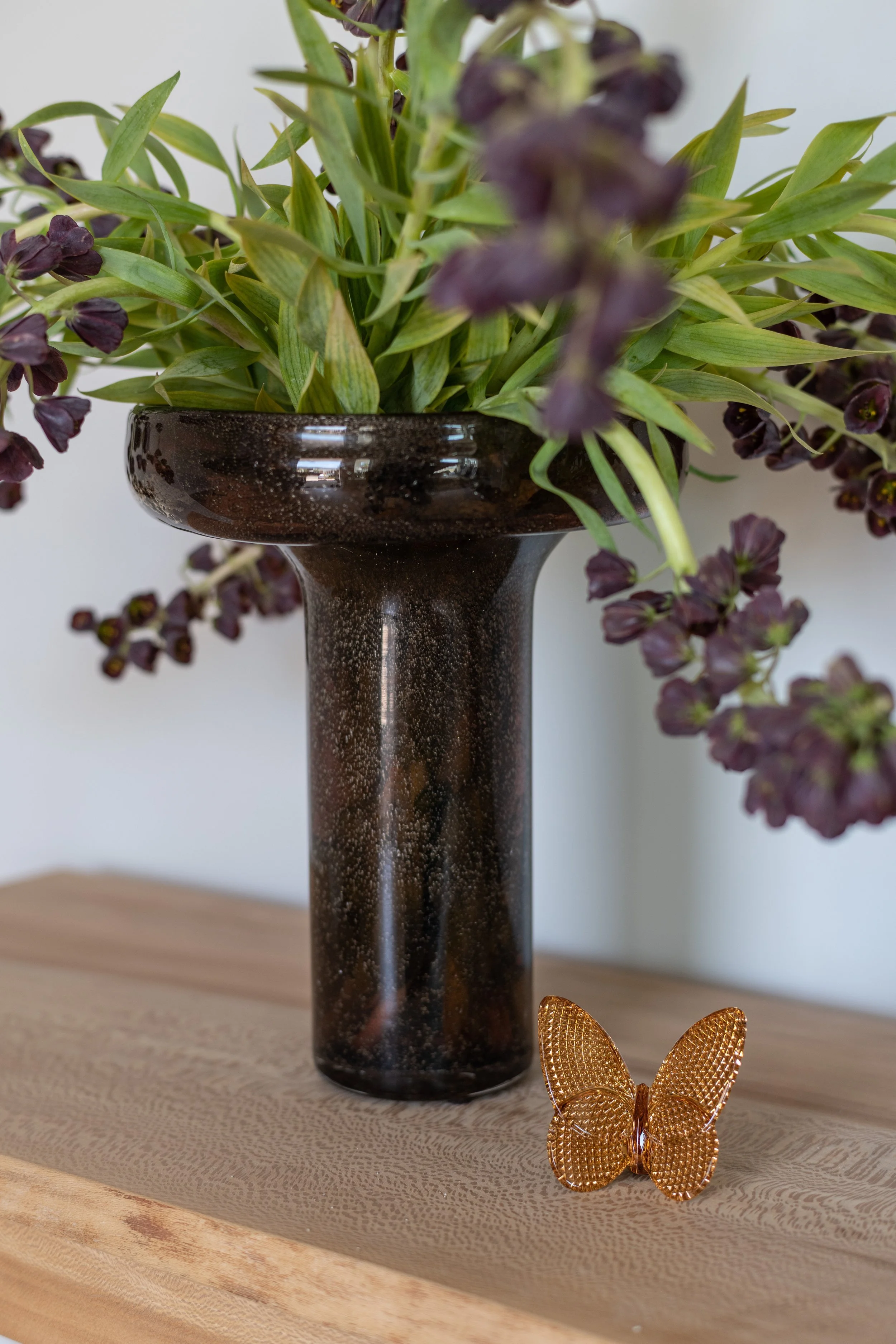 A tall, black speckled vase holding purple flowers and green foliage on a wooden surface, with a decorative gold butterfly ornament in front. Interior design by Sarah Barnard. 