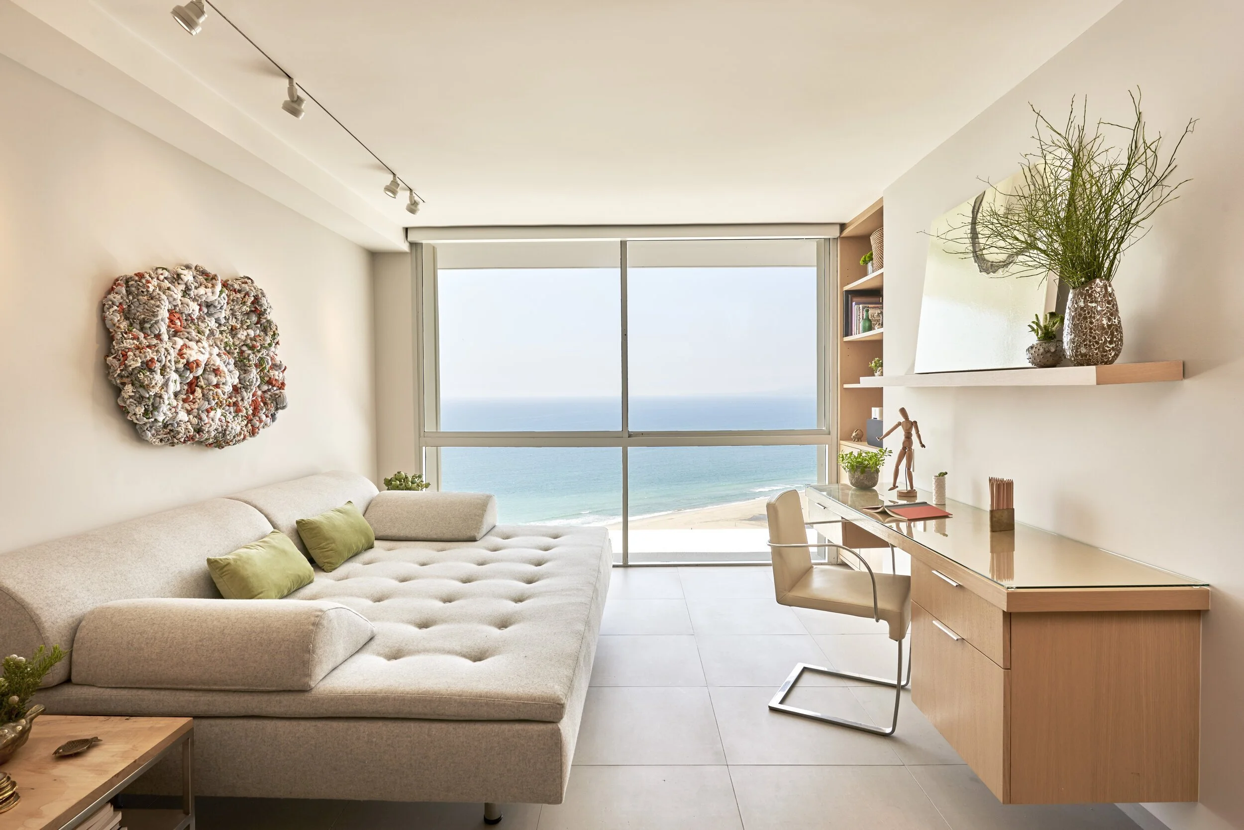 Living room with a large beige sectional sofa, a wooden desk with a beige chair, and a large window view of the ocean and beach. Interior design by Sarah Barnard.
