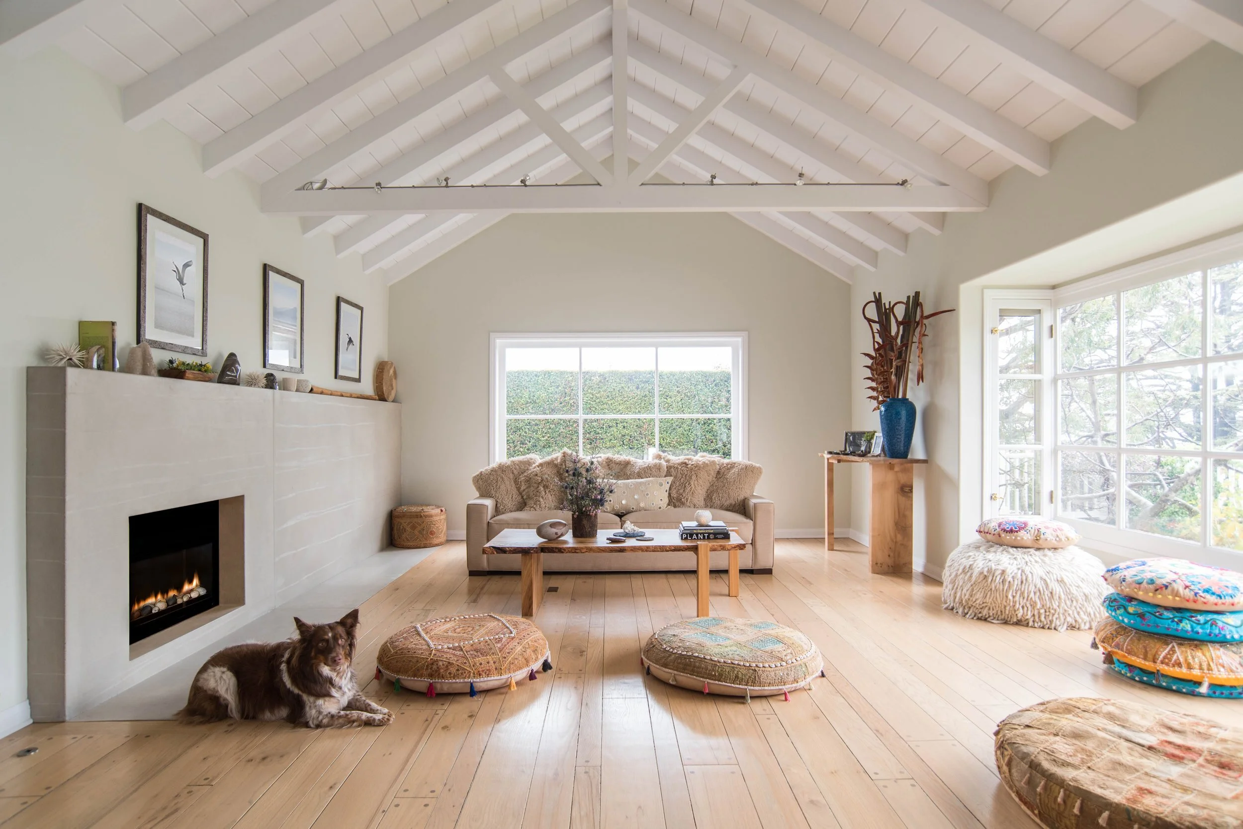 Bright living room with vaulted ceiling, large window, sofa with cushions, fireplace, floor cushions, wooden coffee table, and decorative items. Interior design by Sarah Barnard.