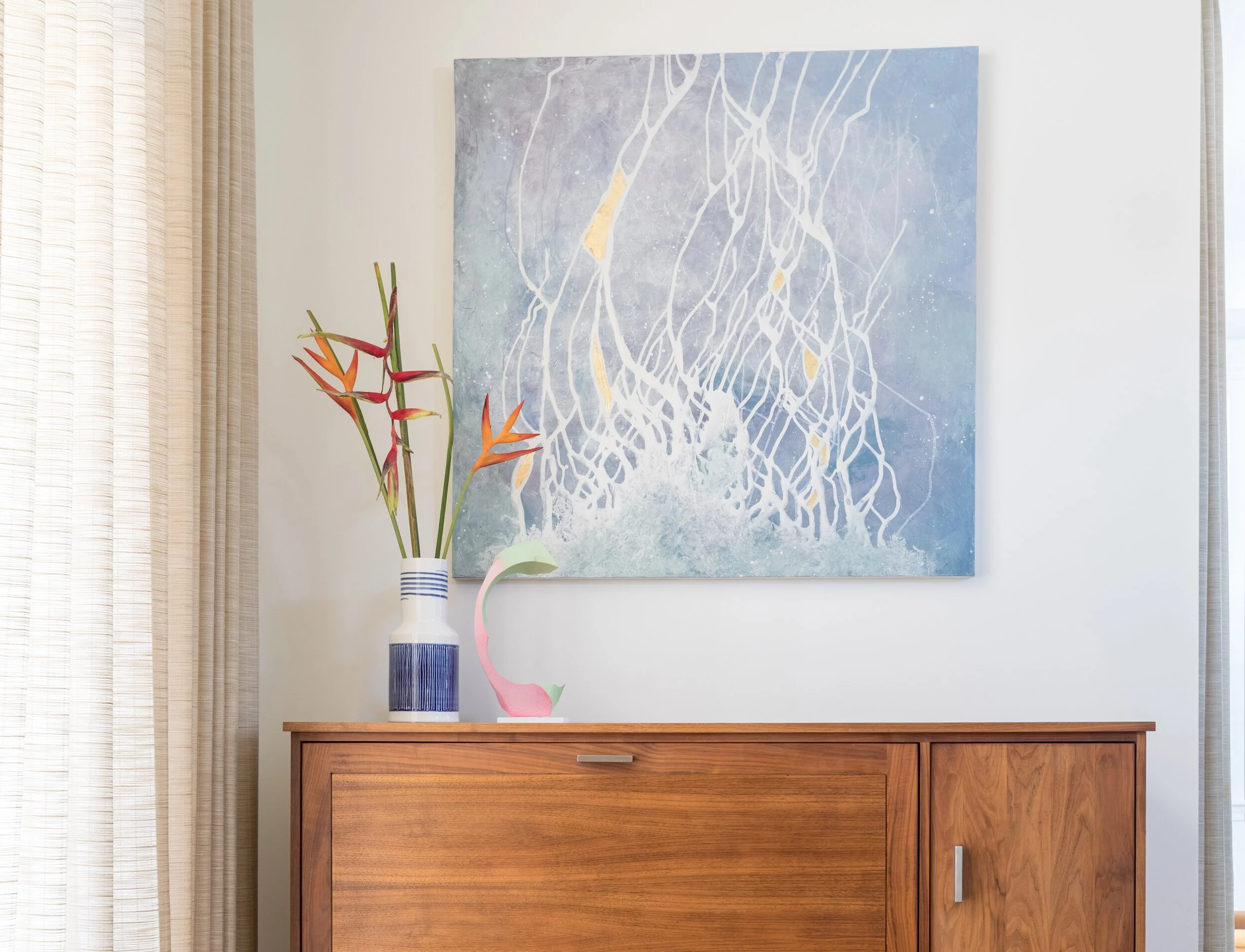 Decorative interior with abstract blue and white painting, a vase with tropical flowers, and a pastel-colored sculpture on a wooden sideboard. Interior design by Sarah Barnard. 