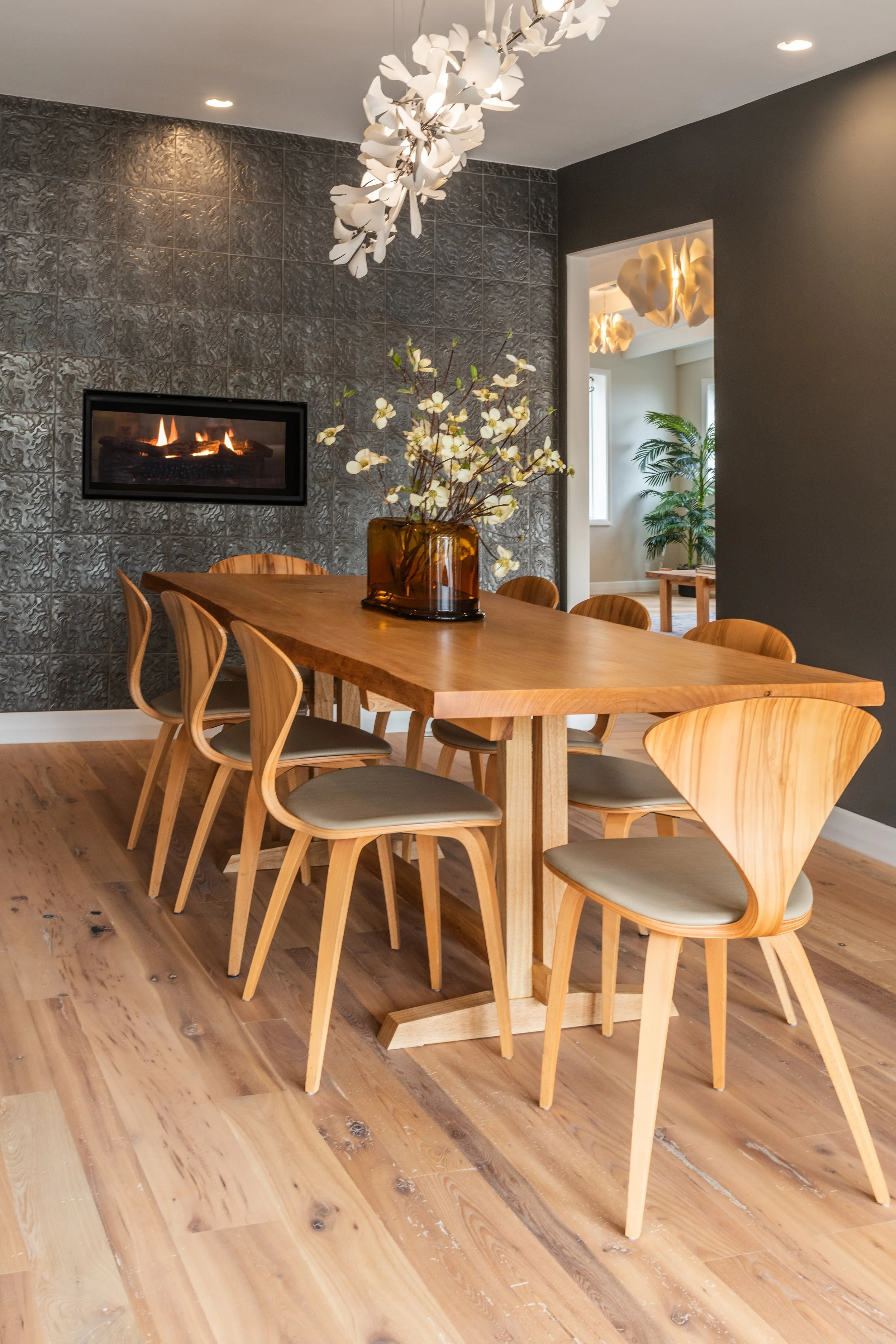 A modern dining room with a wooden table and eight matching wooden chairs, a tall amber glass vase with white flowers on the table, a textured dark gray wall with a mounted fireplace. Interior design by Sarah Barnard. 