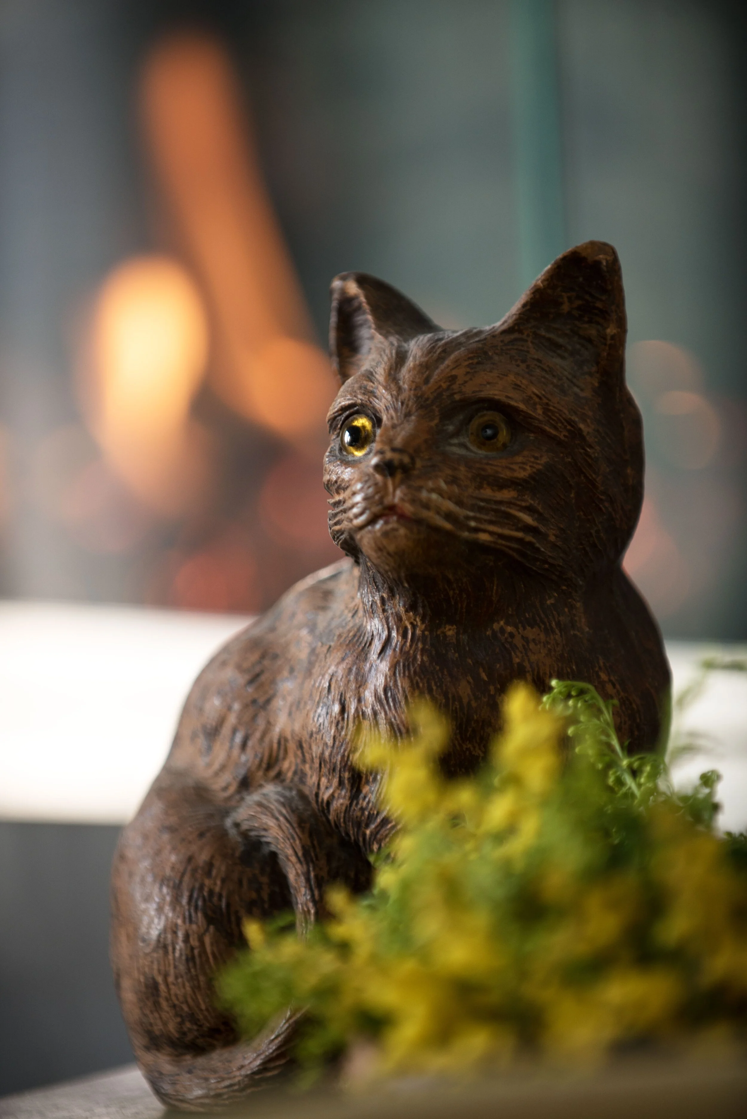 A detailed wooden sculpture of a cat with realistic eyes, placed among yellow flowers, with a blurred background featuring warm and cool lights. Interior design by Sarah Barnard. 