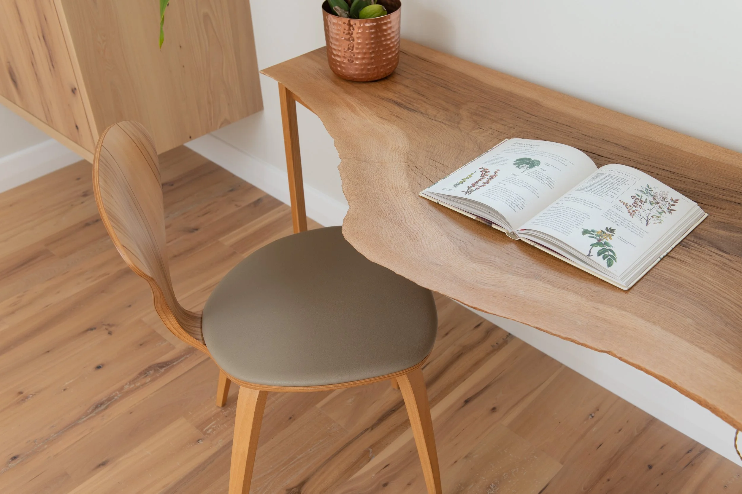 A wooden desk with a natural edge, an open botanical book, and a small potted plant in a copper-colored container. A wooden chair with a cushioned seat is positioned at the desk. Interior design by Sarah Barnard. 