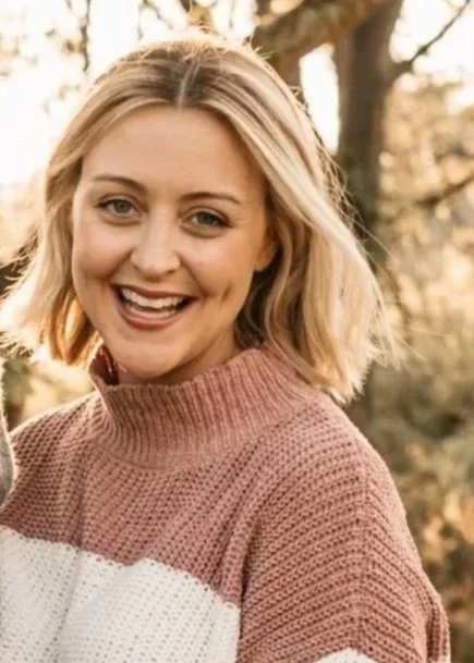 A woman with shoulder-length blonde hair smiling outdoors, wearing a pink and white sweater.