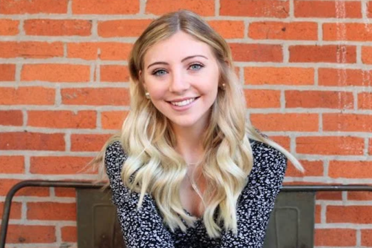 A young woman with long blonde hair, smiling, wearing a black and white polka dot top, sitting in front of a brick wall.