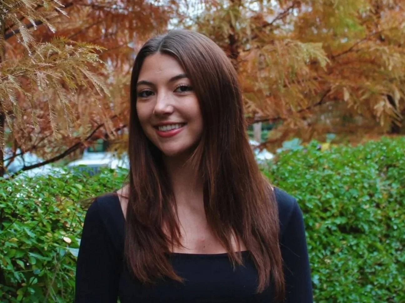A young woman with long brown hair smiling outdoors in front of orange autumn trees and green bushes.