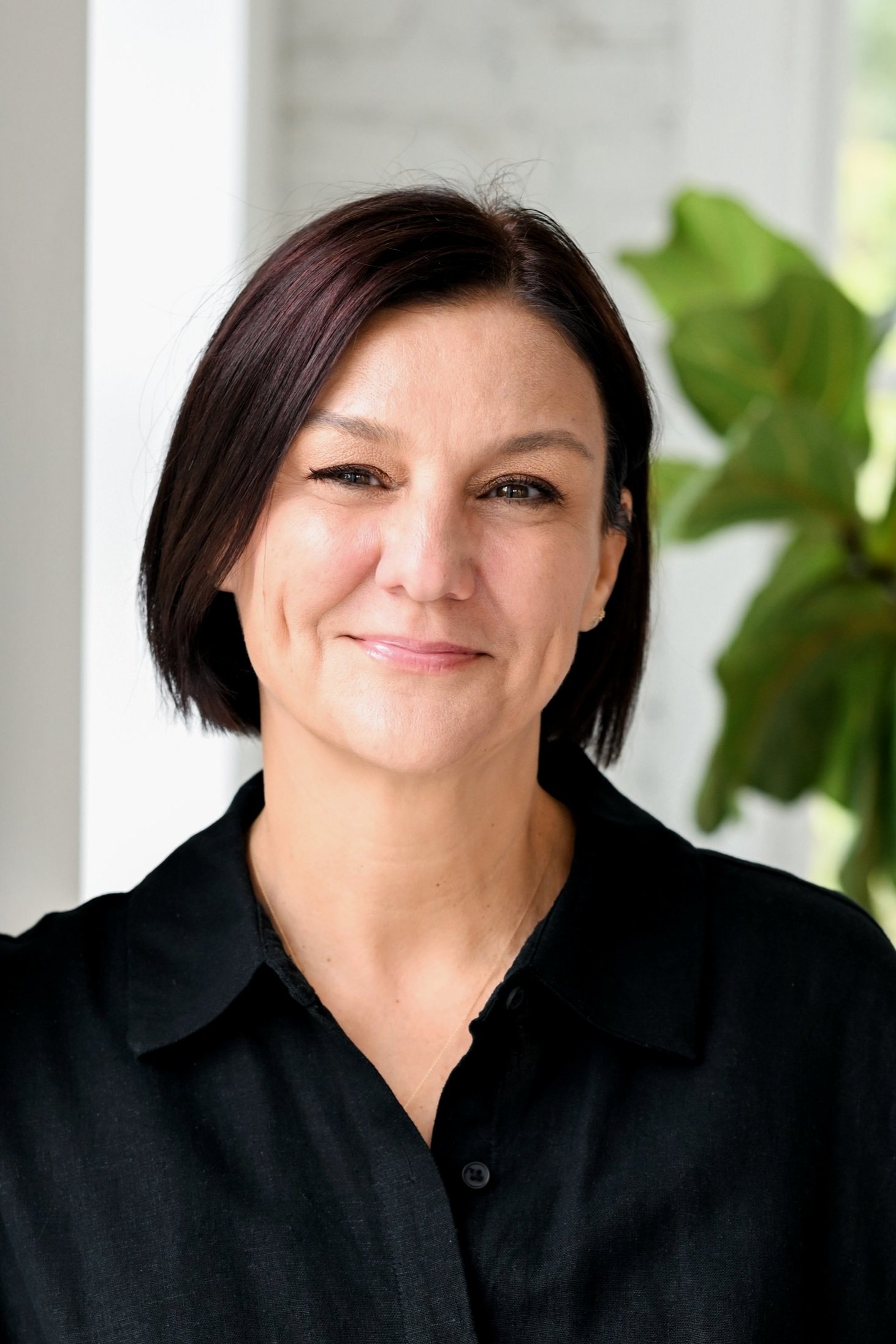 A woman with short dark hair, wearing a black shirt, smiling and winking at the camera. Behind her is a blurred background with a large green plant.