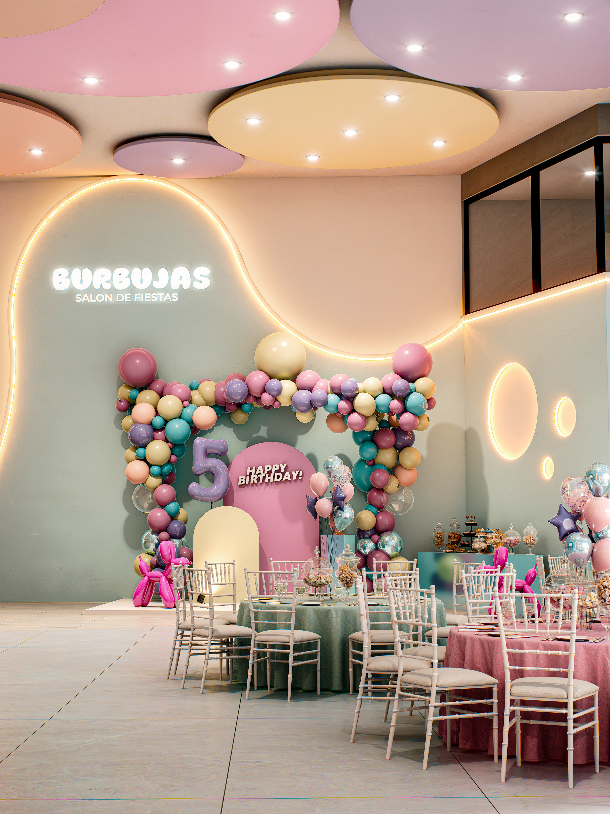 Decoración de fiesta con globos de colores pasteles, cartel de cumpleaños que dice 'Feliz cumpleaños', mesas con manteles pastel y sillas blancas, paredes con luces de neón y decoración de luna y burbujas en tonos suaves.