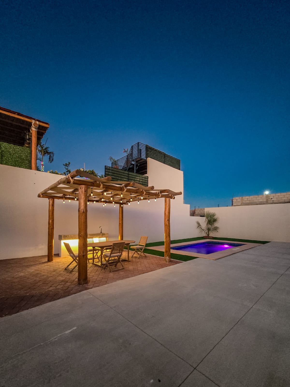 Área exterior de una casa con una piscina iluminada, una pergola de madera con una mesa y sillas, paredes blancas, una pequeña planta en la esquina y un cielo nocturno despejado.