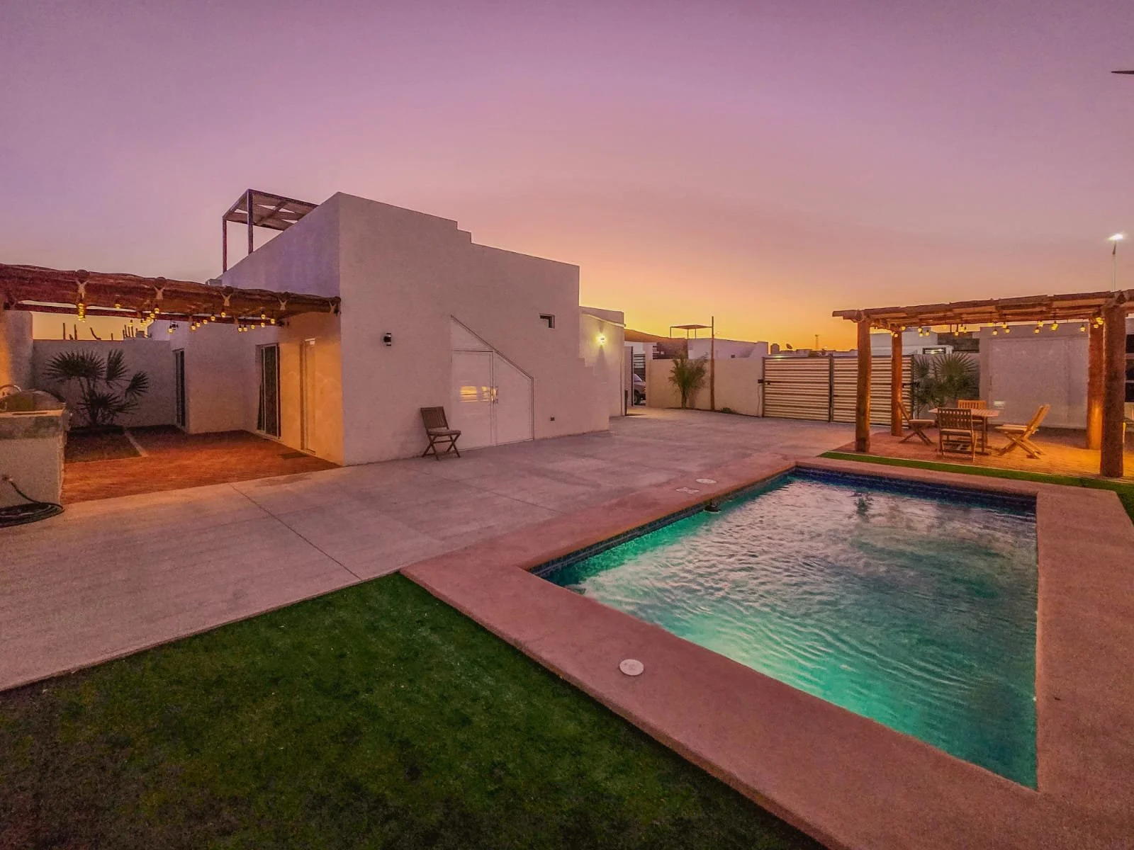 Cerca de la piscina en un patio con áreas pavimentadas y césped, con una estructura de pérgola de madera con iluminación y mobiliario de comedor, al atardecer con un cielo anaranjado y morado.