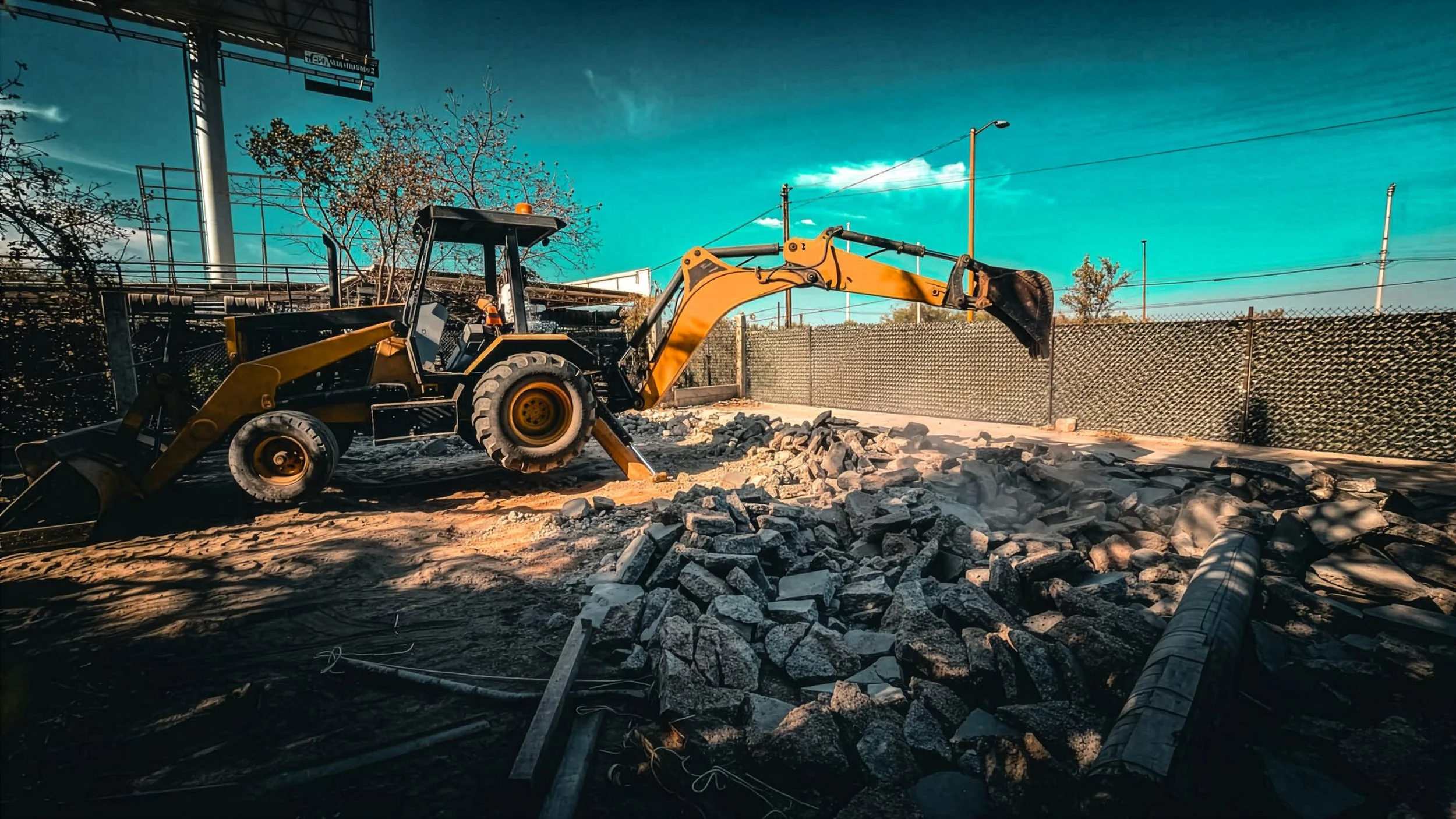 Retroexcavadora amarilla en un sitio de construcción con escombros y tuberías, bajo un cielo azul. Muestra el inicio del proceso de construcción.