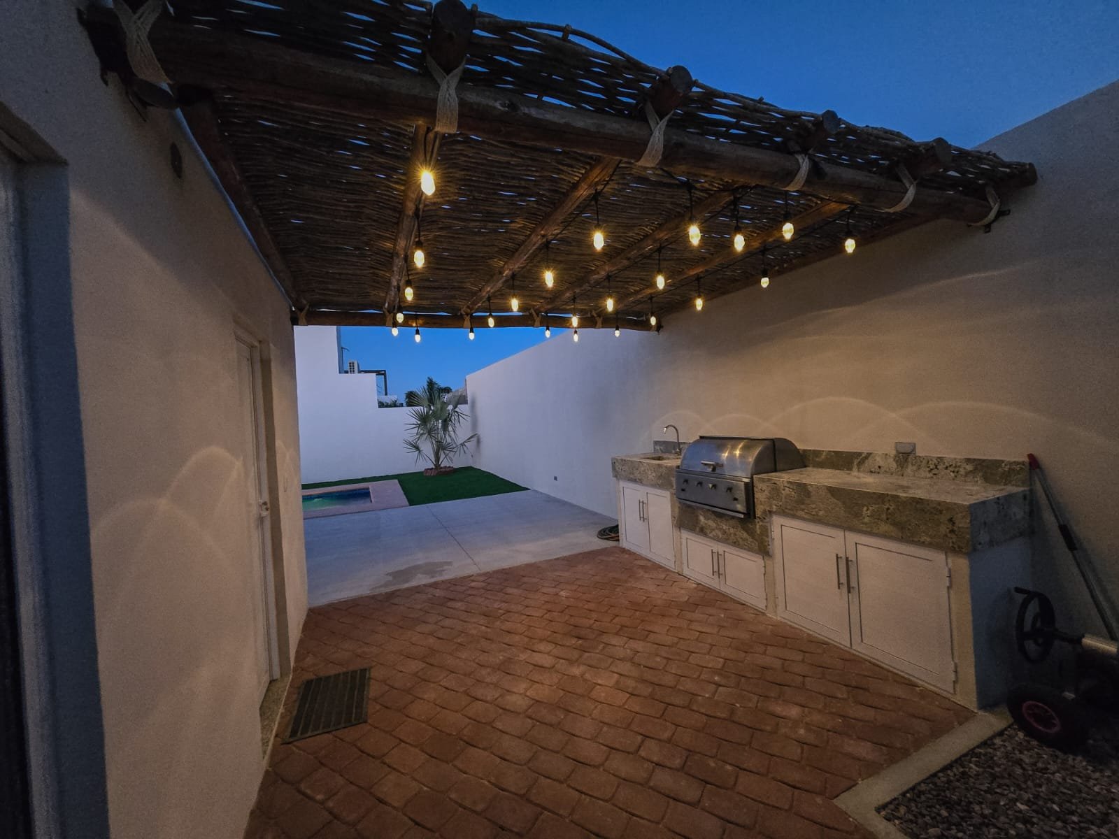 Área de barbacoa con paredes blancas, techo de madera con luces colgantes, cocina con barra de granito y fregadero, piso de ladrillo y cerca de una piscina y plantas.
