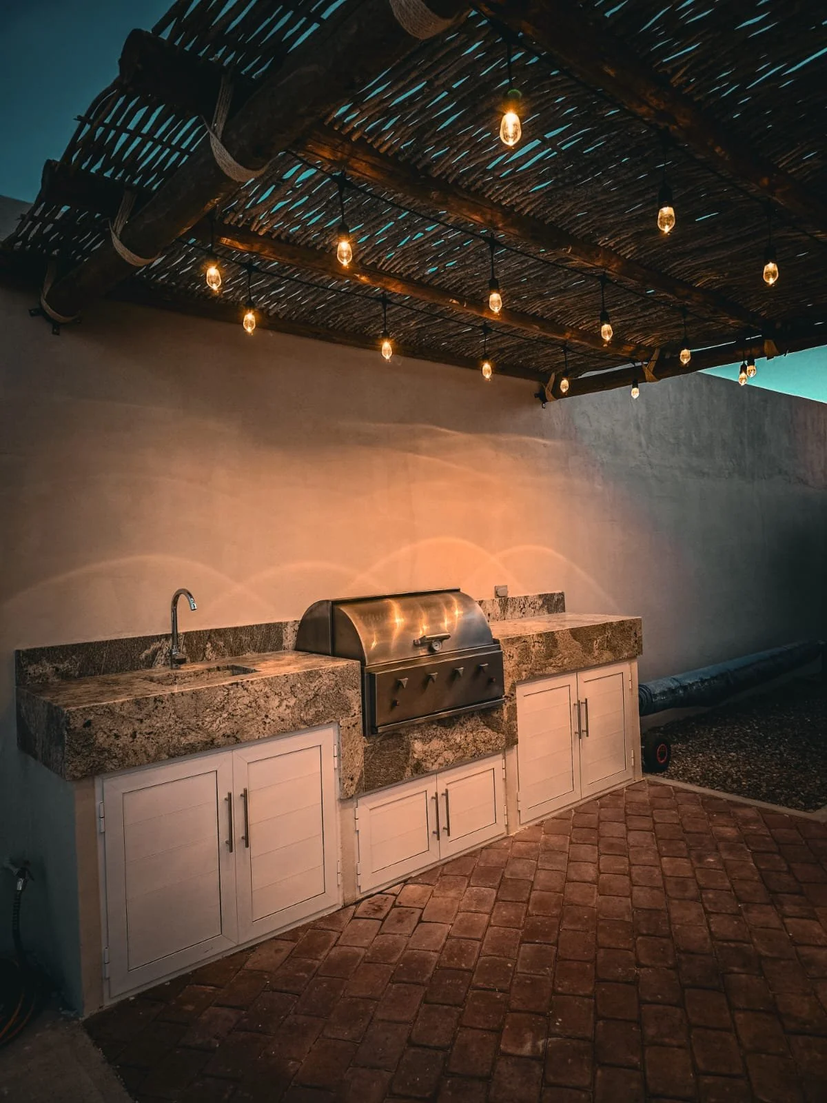 Área de cocina exterior con encimera de granito, fregadero y parrilla de metal, bajo un techo de madera con luces decorativas, suelo de ladrillos y pared de color claro.