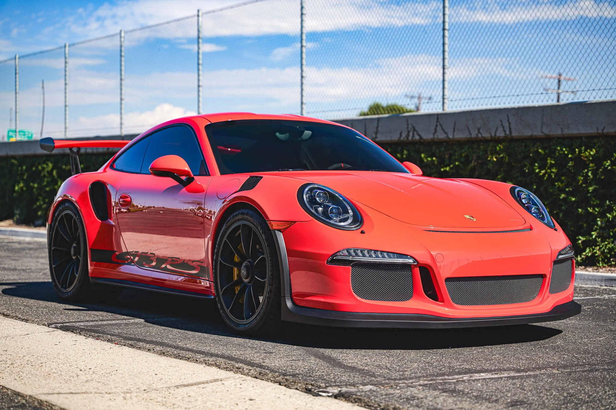 Red Porsche 911 GT3 RS sports car parked on a street with a chain-link fence and blue sky in the background.