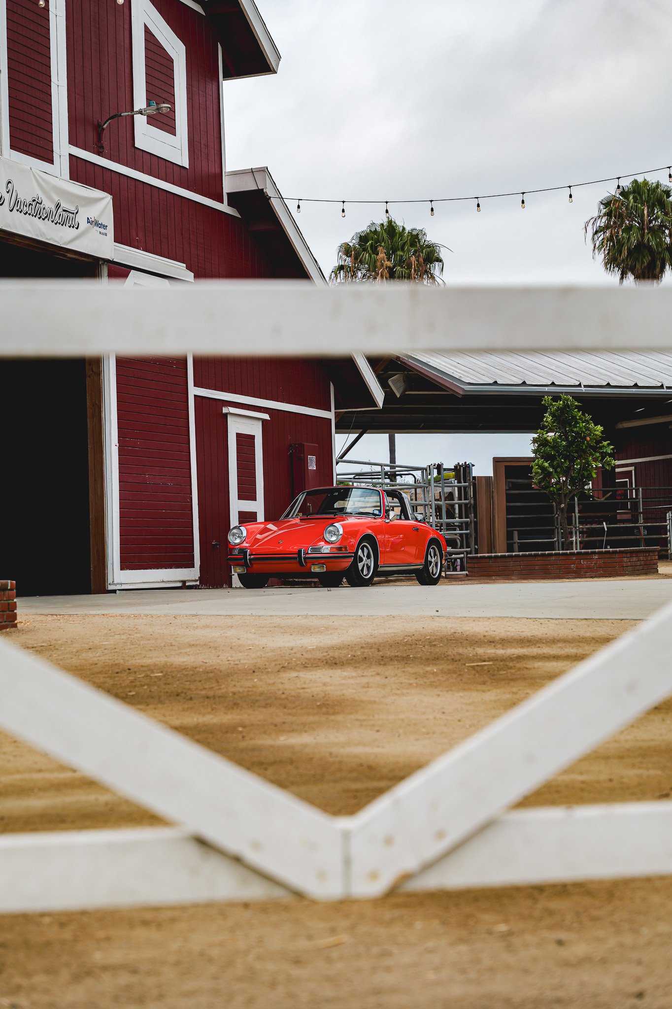 Red vintage Porsche car parked near a red barn with a metal roof, seen through a white lattice fence with a sidewalk and some trees in the background.