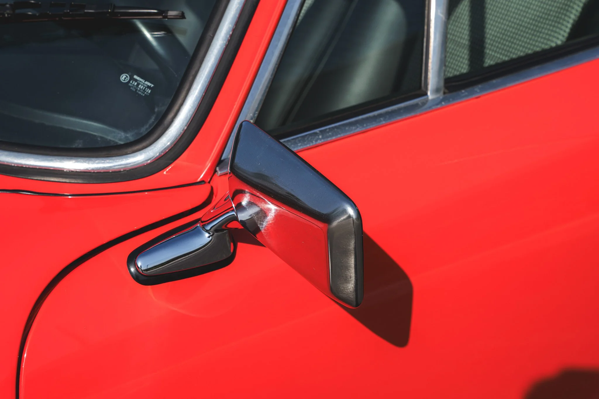 Close-up image of a vintage red car with a side mirror and windshield visible, showing part of the vehicle's front and side.