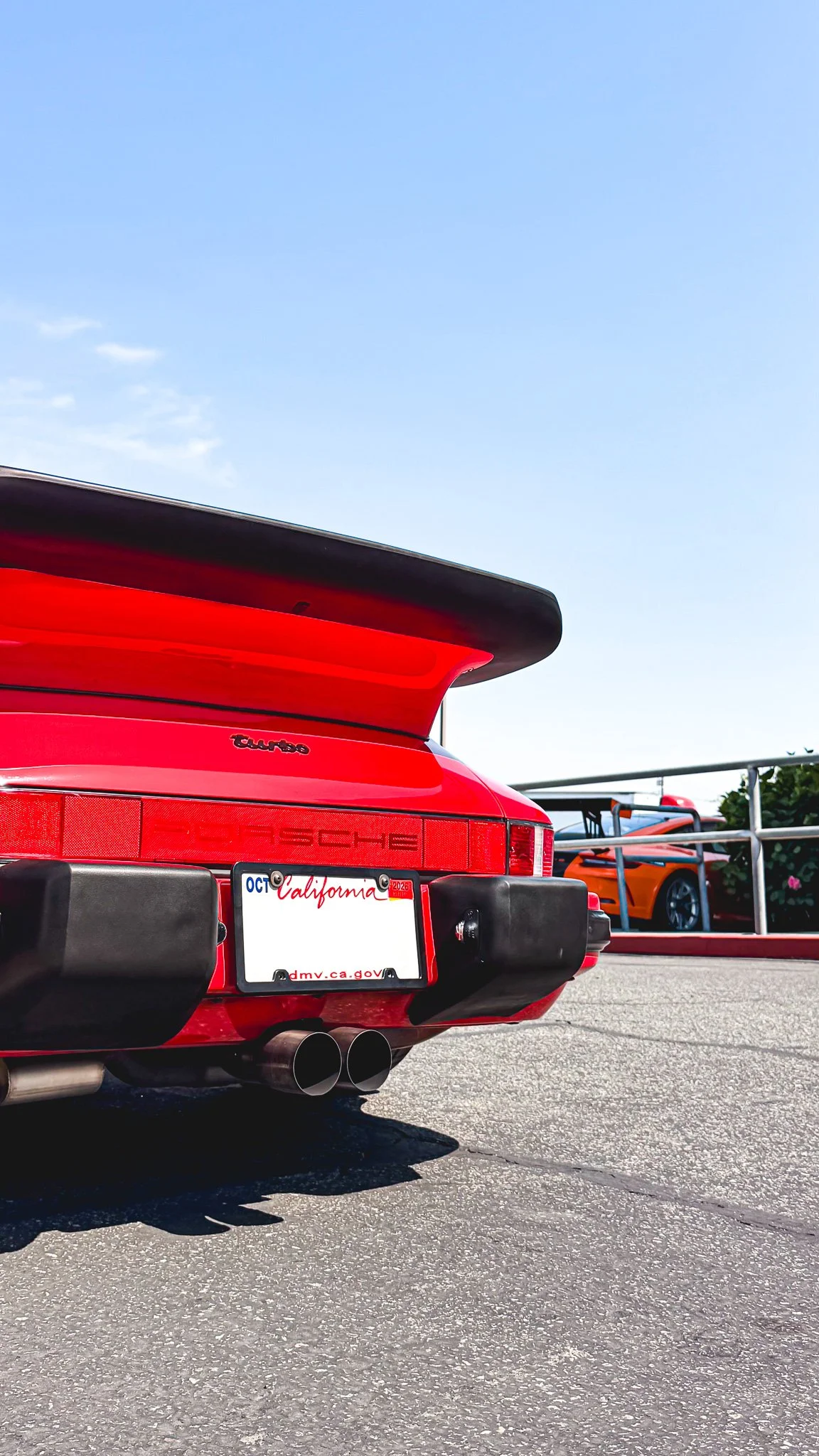 The back of a red Porsche sports car with a large rear wing, dual exhaust pipes, and California license plate, parked in a lot with a few other sports cars and a clear blue sky above.