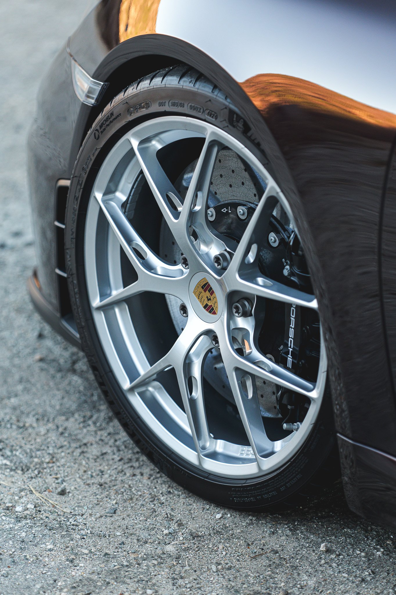 Close-up of a black Porsche car wheel with car logo, silver alloy rim, and Michelin tire, parked on a gravel surface.
