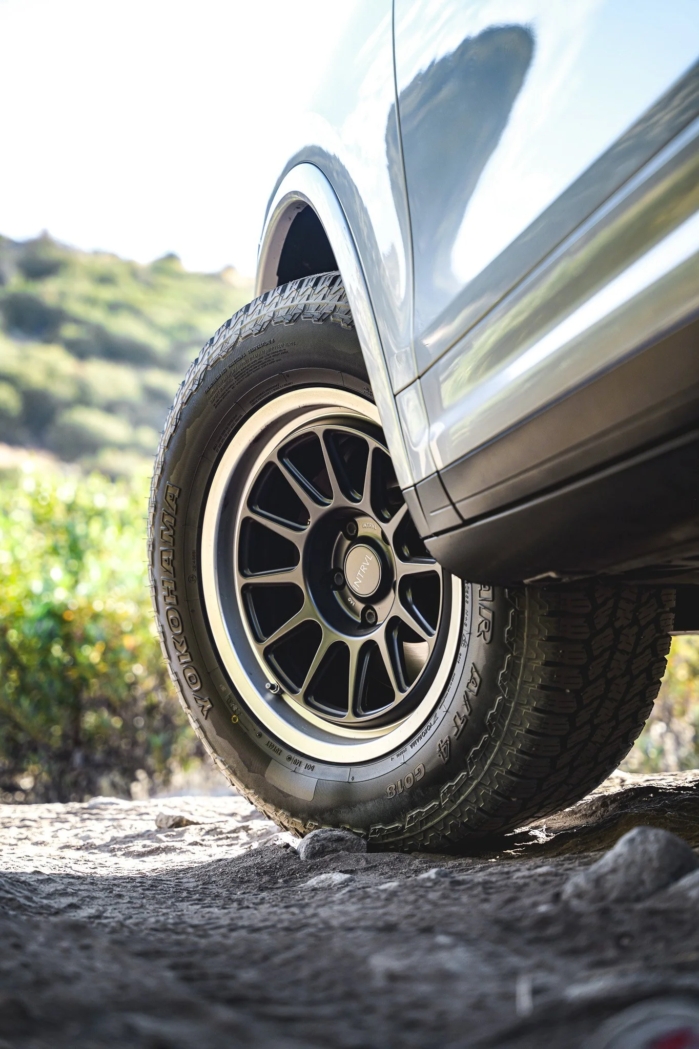 Close-up of a black off-road tire and wheel of an SUV vehicle driving on uneven rocky terrain, with a blurred natural landscape in the background.