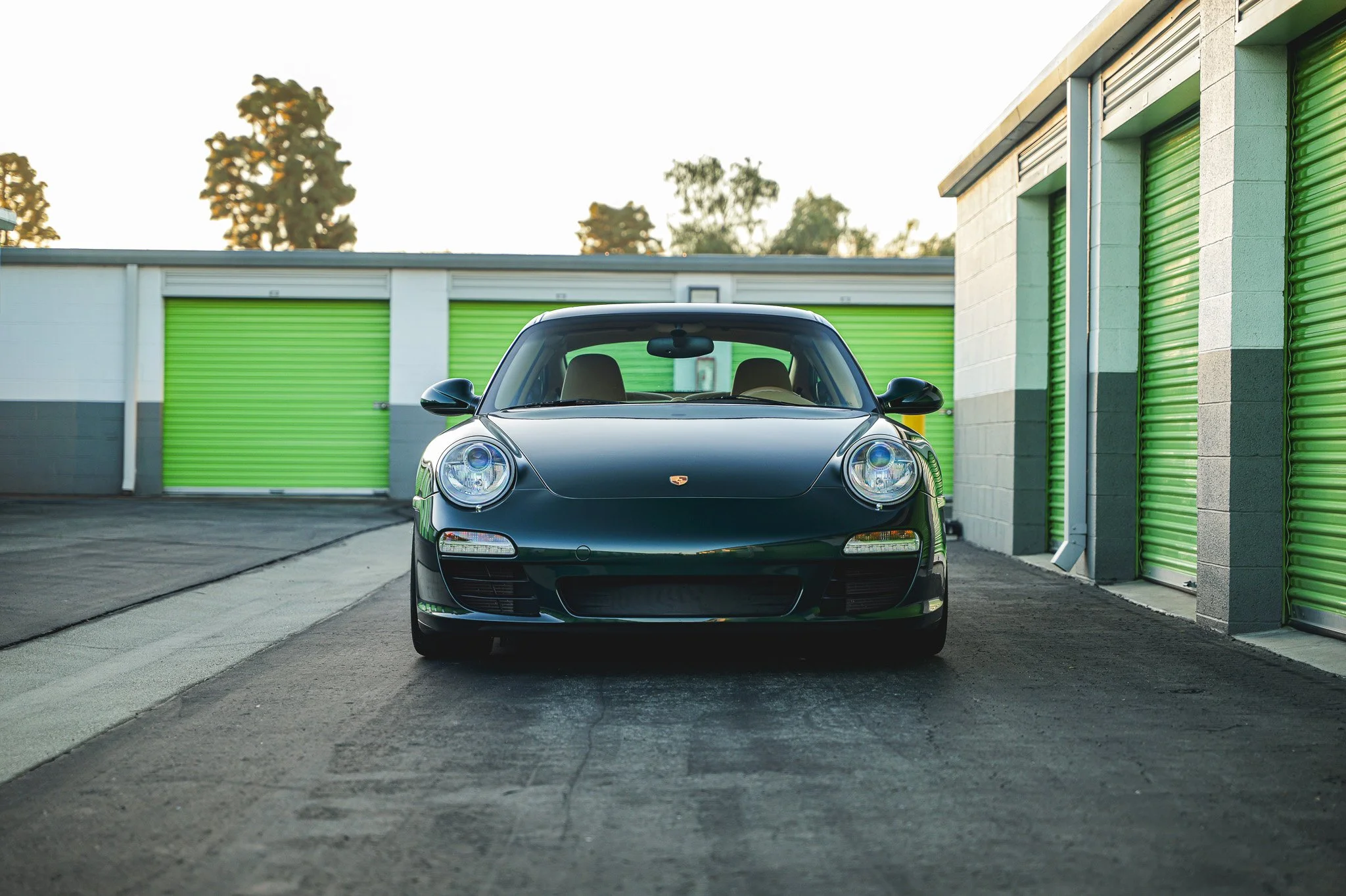 Black Porsche sports car parked outside storage units with green roll-up doors in the background.