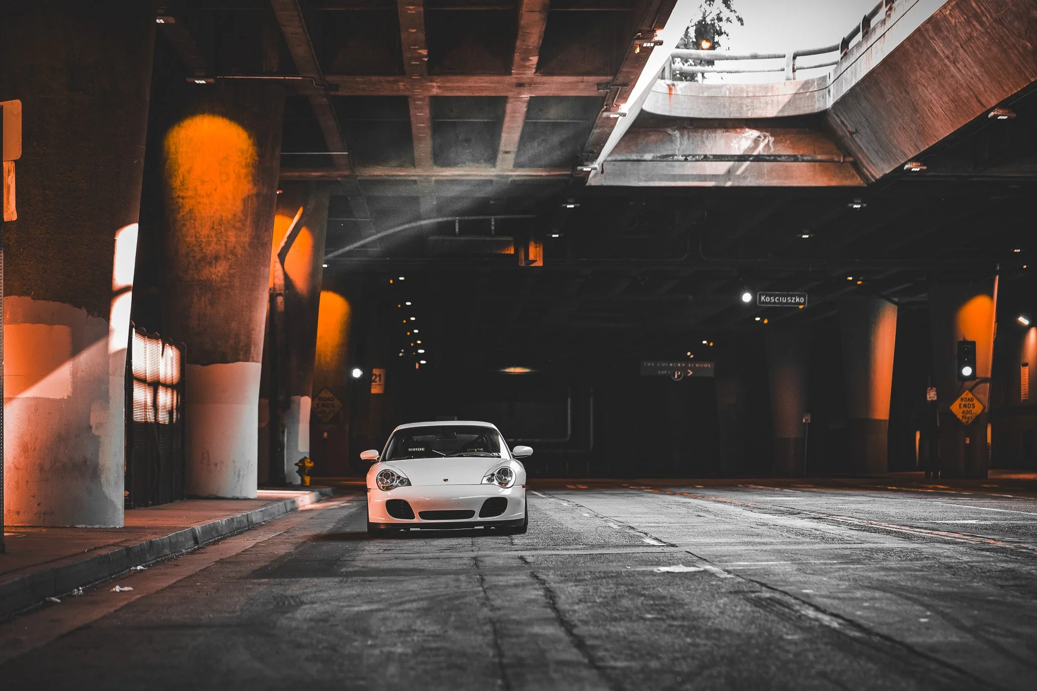 A white sports car parked underneath an overpass in an urban area, illuminated by streetlights with reflections on the wet pavement.