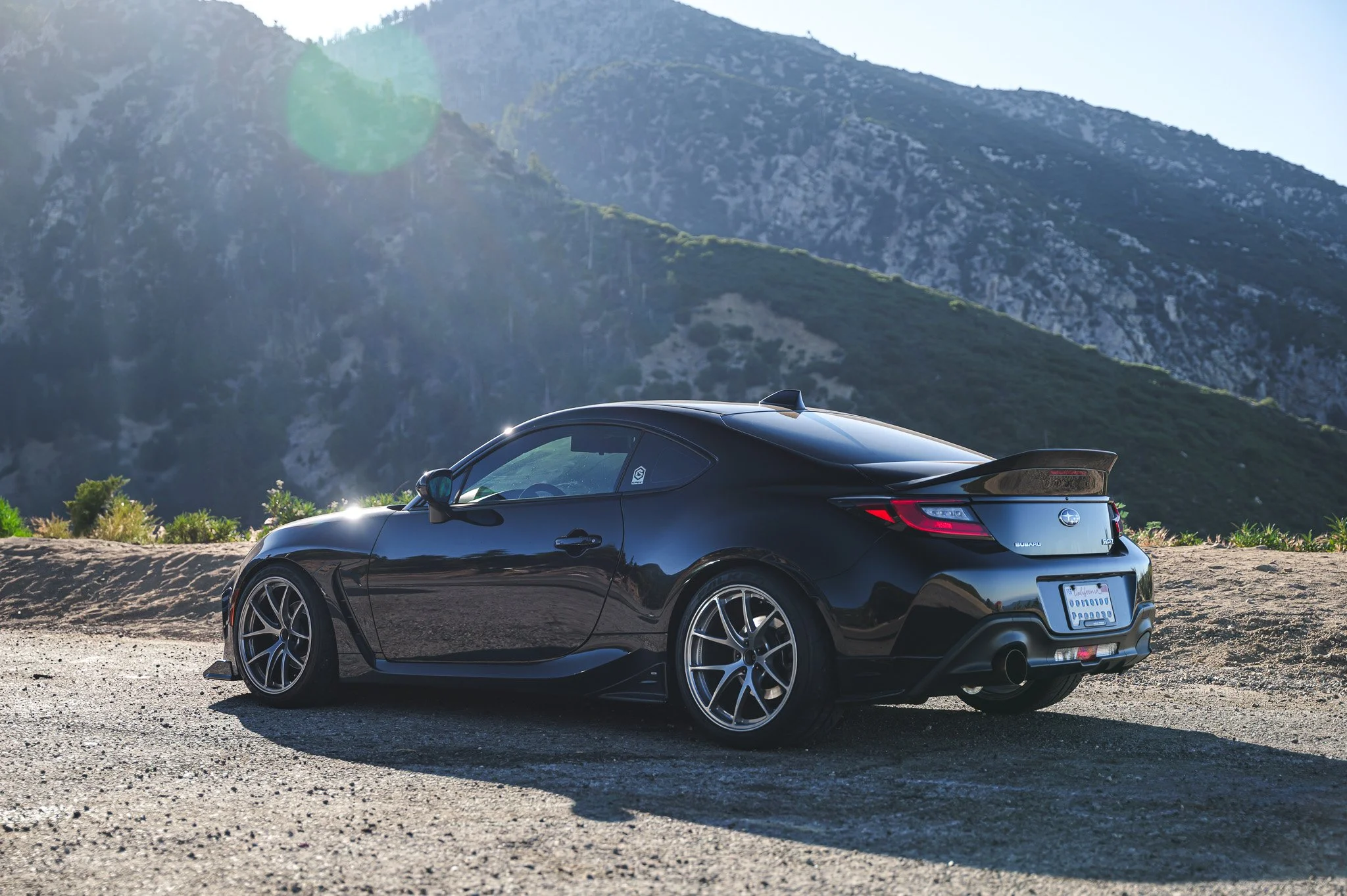 Black Subaru sports car parked on a gravel roadside with mountainous landscape in the background under bright, clear sky.