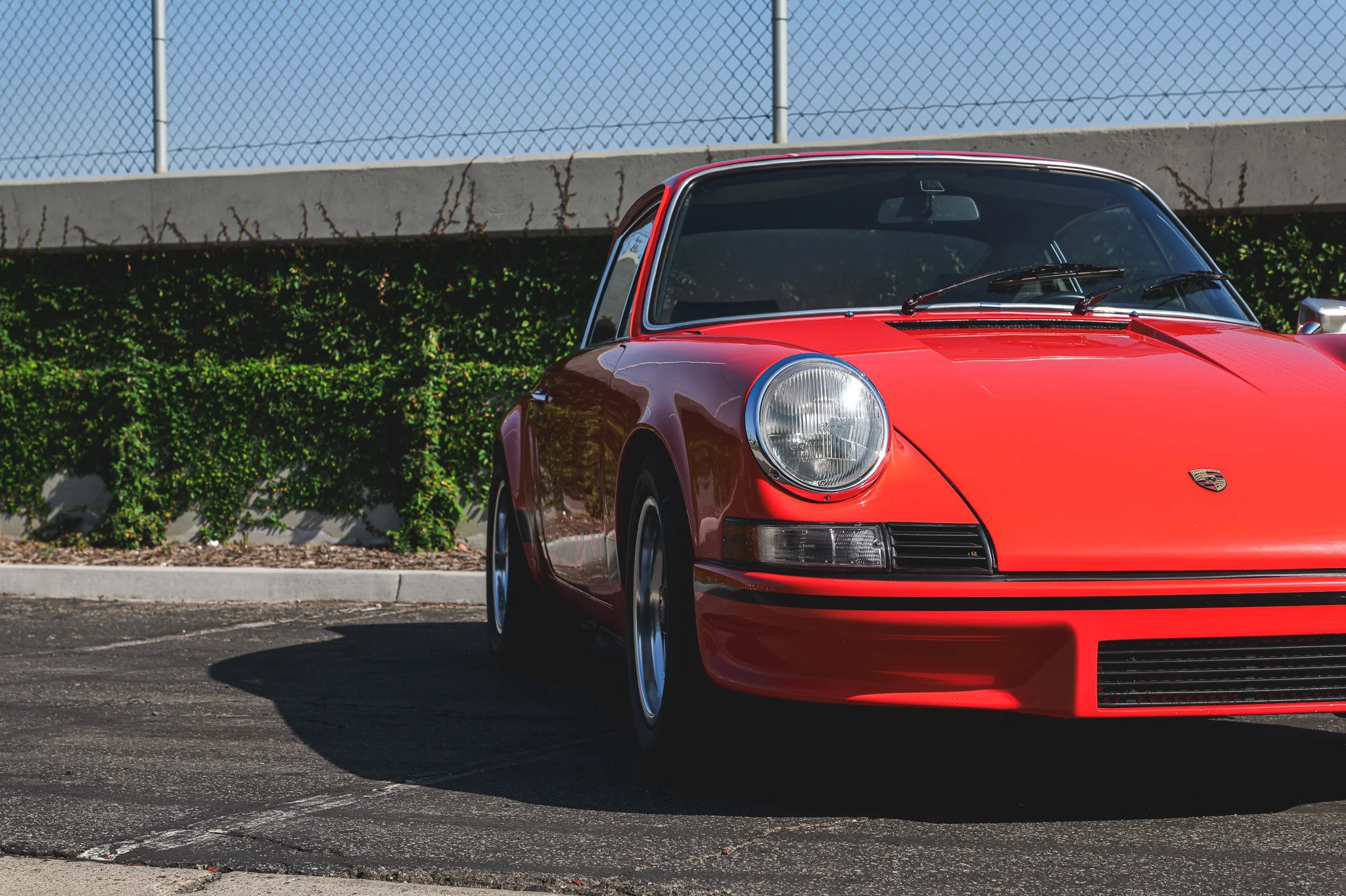 A red classic Porsche sports car parked in a parking lot with a green hedge and a chain-link fence in the background.