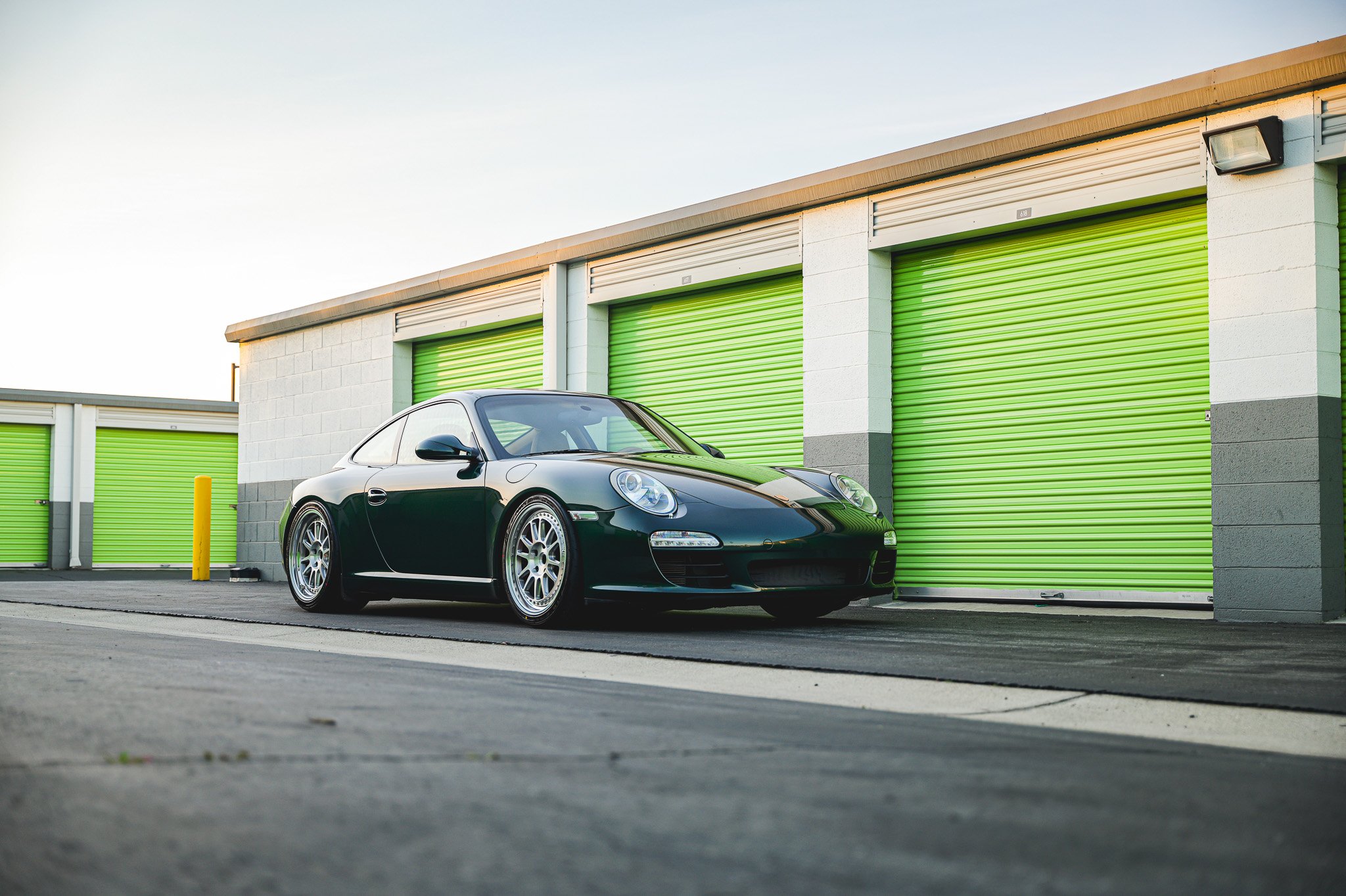 A black Porsche sports car parked in front of self-storage units with green roll-up doors.