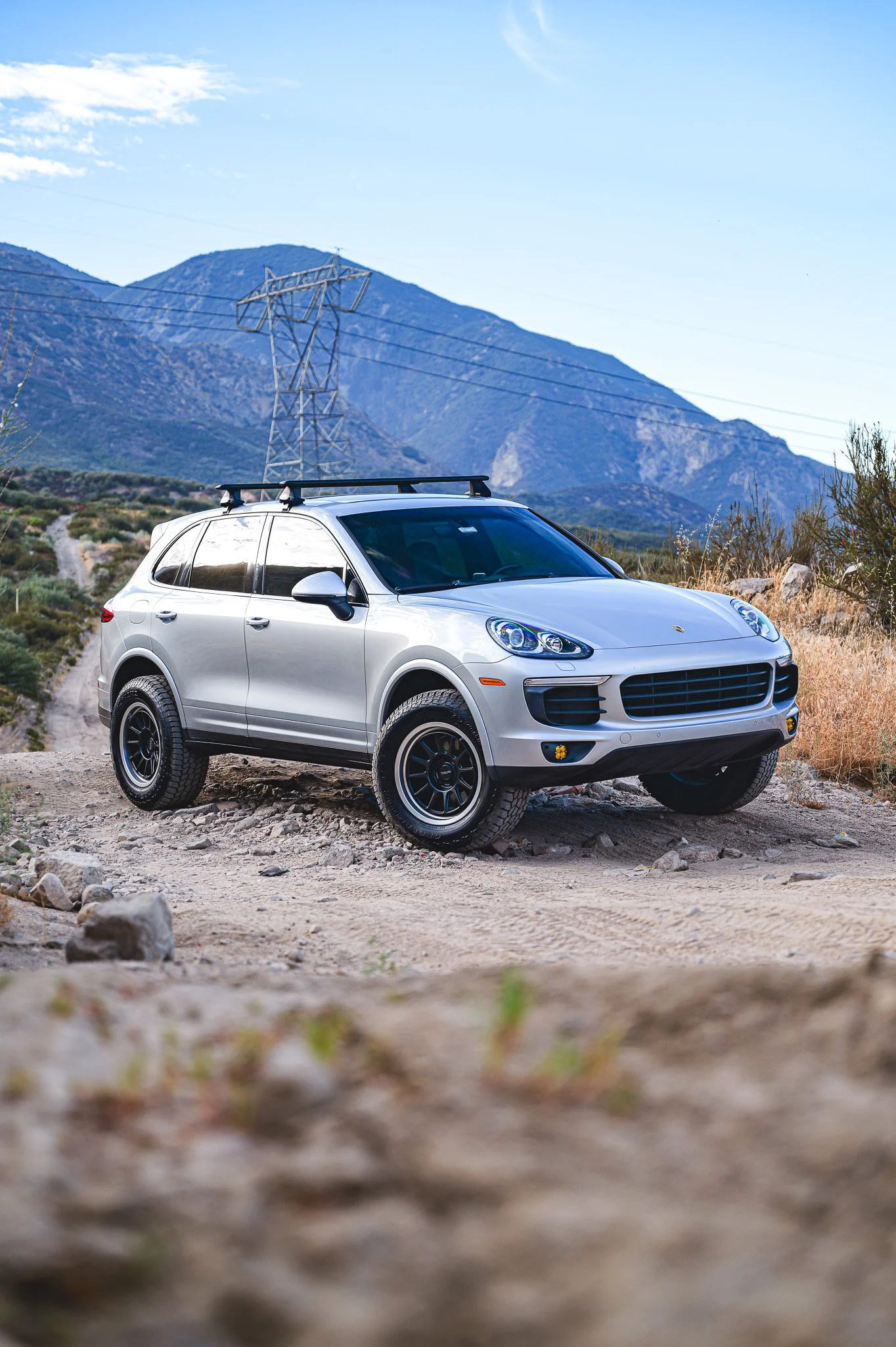 A silver Porsche SUV parked on a rocky dirt road with mountains in the background and power lines overhead.