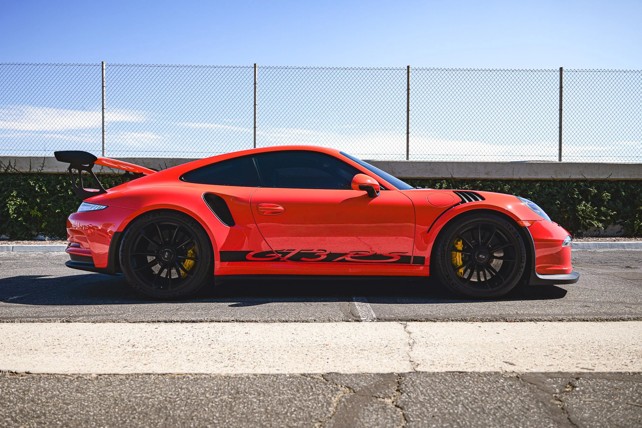 Red sports car with black wheels and a rear spoiler parked on a street in front of a chain-link fence and clear blue sky.