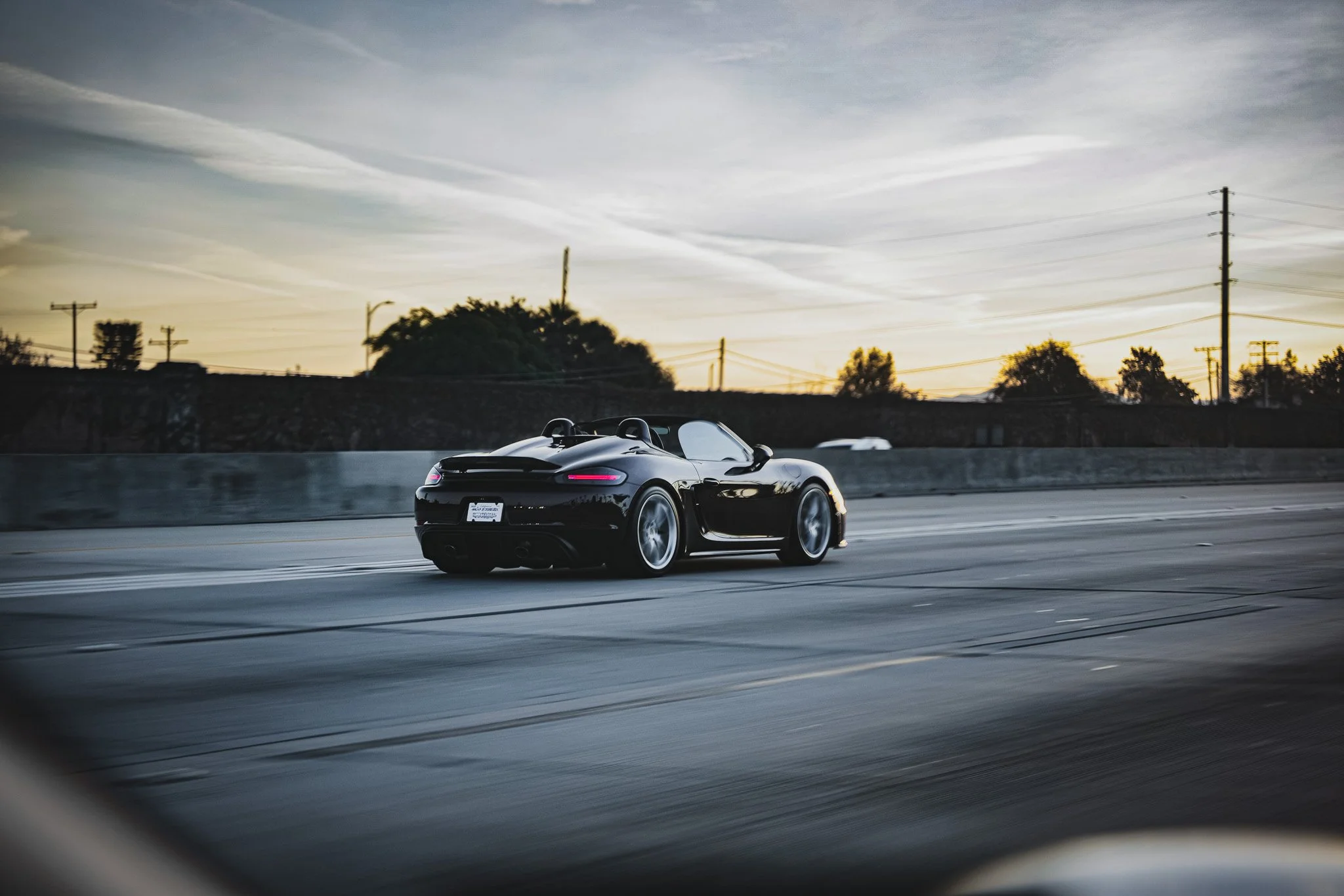 A black convertible sports car driving on a highway at sunset with trees and utility poles in the background.