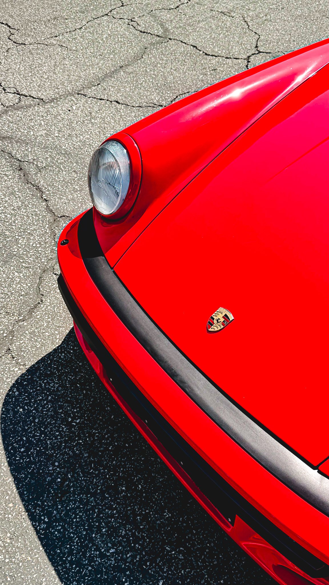 Close-up of the front of a red Porsche sports car, showing the headlight, hood, and Porsche emblem, parked on asphalt.