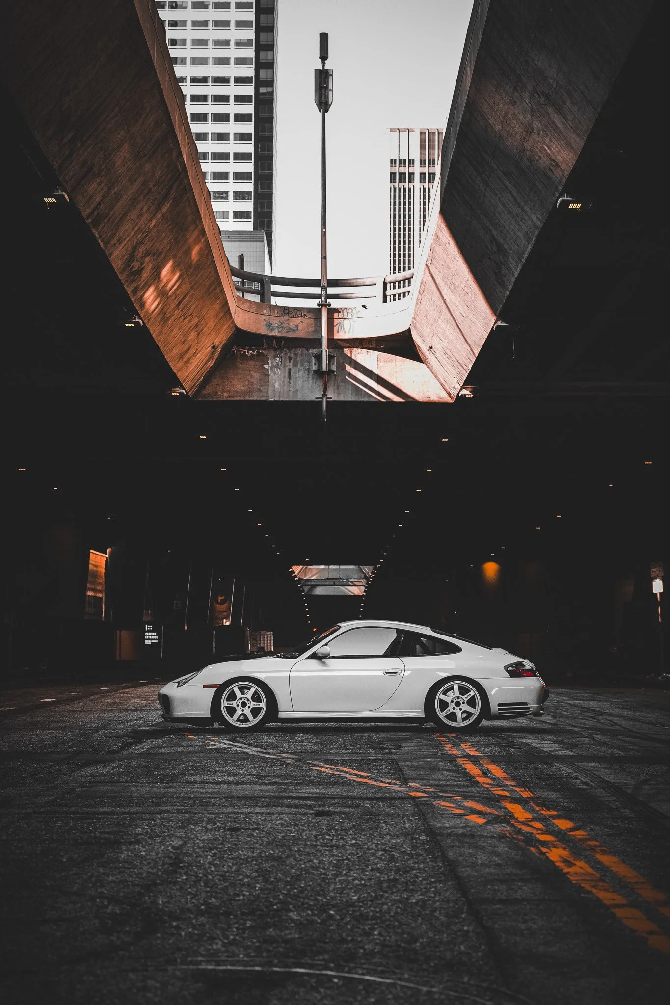 A white sports car parked in an urban parking lot beneath an overpass with tall buildings in the background. The scene is moody and dimly lit.