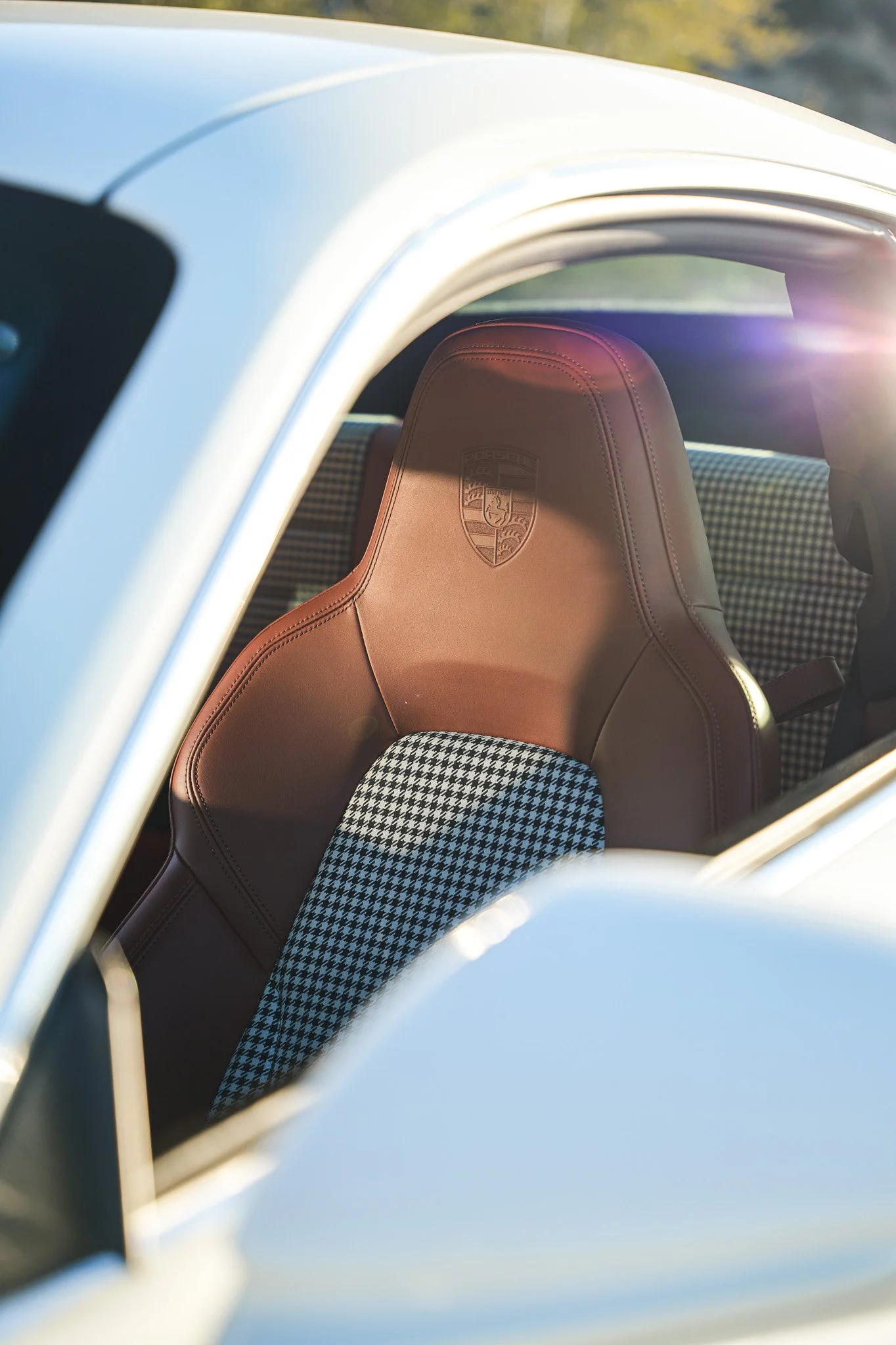 Close-up view of the interior of a car, featuring a brown leather seat with the Porsche logo embossed on the headrest, and houndstooth-patterned upholstery on the seat back. The car's exterior appears to be white, with sunlight shining into the windo