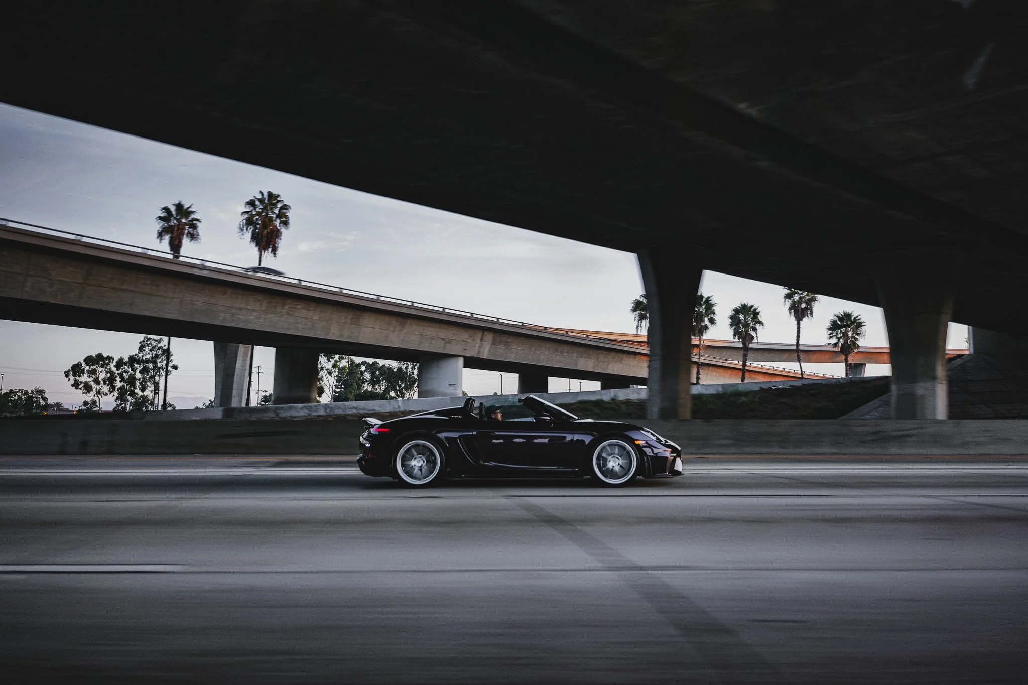 A black convertible sports car driving on a highway under an overpass with palm trees in the background.