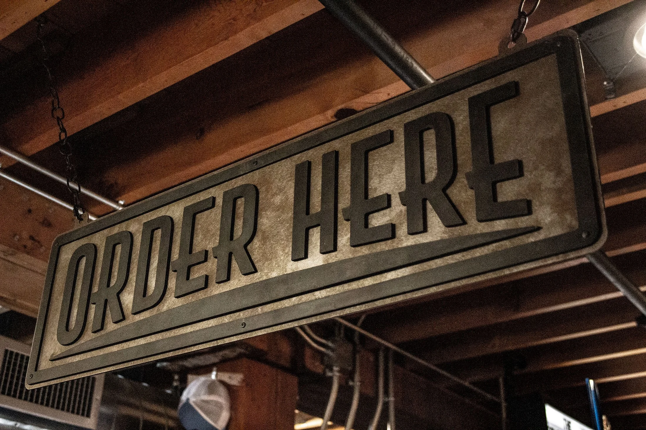 A rectangular metal sign with black raised letters reading 'ORDER HERE' hanging from a chain inside a wooden building or restaurant.